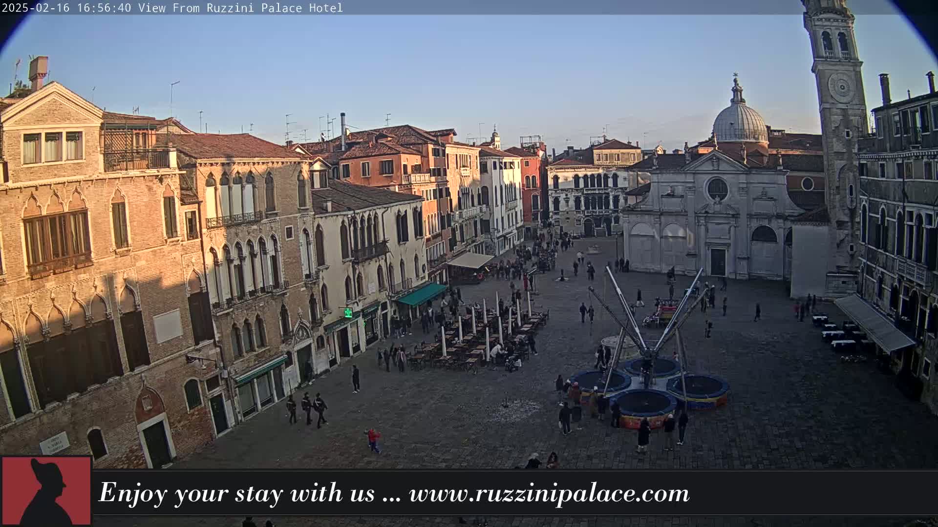A clear, sunny day reveals a Venetian piazza with several buildings, outdoor seating, and a small inflatable trampoline in the center.