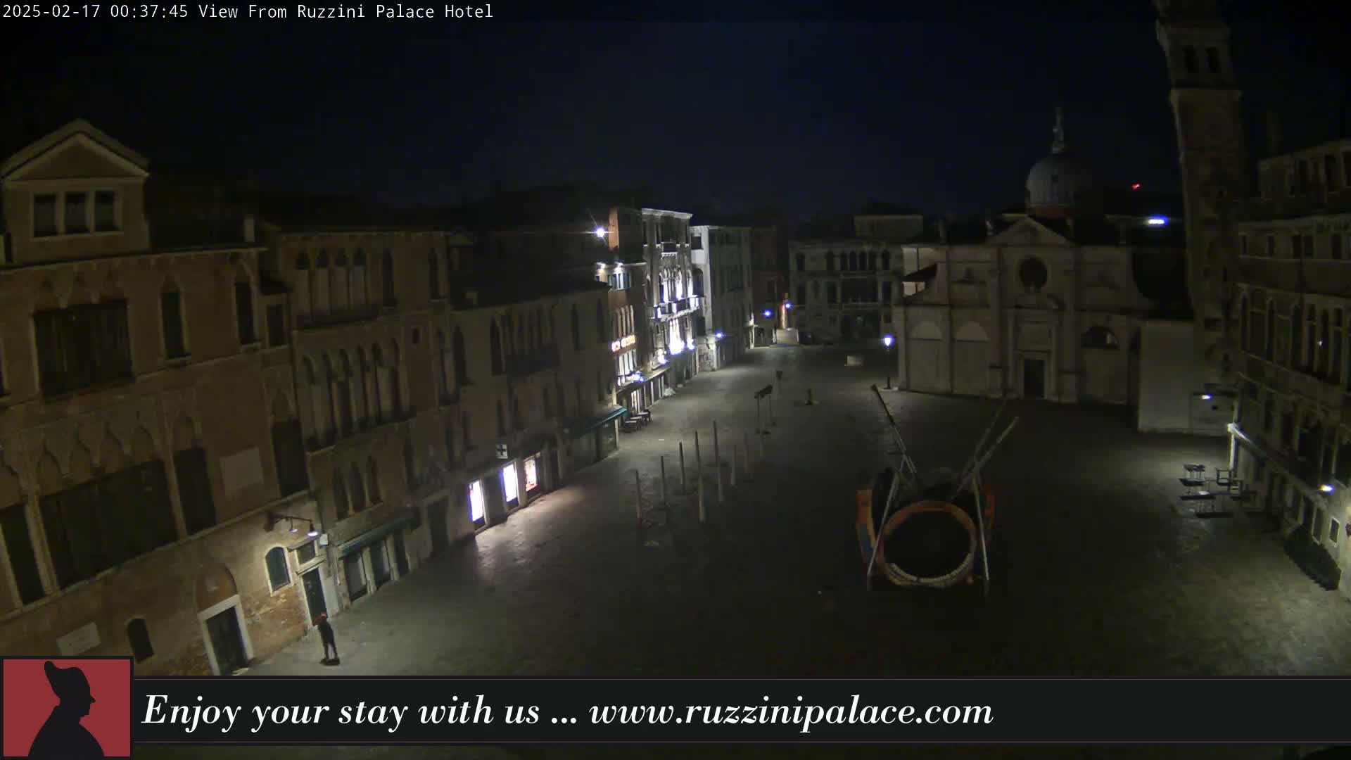 A nighttime view of a mostly empty, cobblestone square in Venice, Italy, surrounded by old buildings, with a few lights illuminating the scene.
