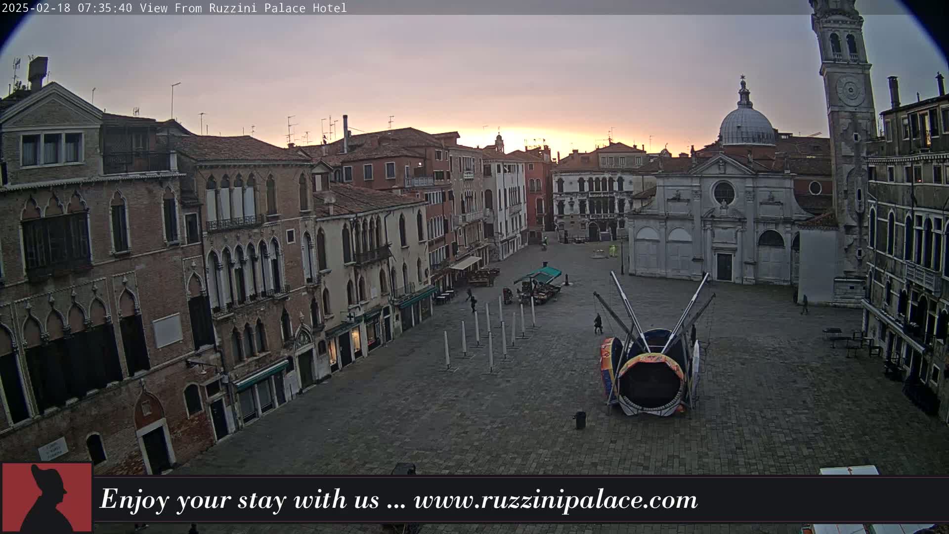 A large, empty plaza in Venice, Italy, is seen at sunrise under a cloudy sky, with several buildings surrounding it and a large circular structure in the center.