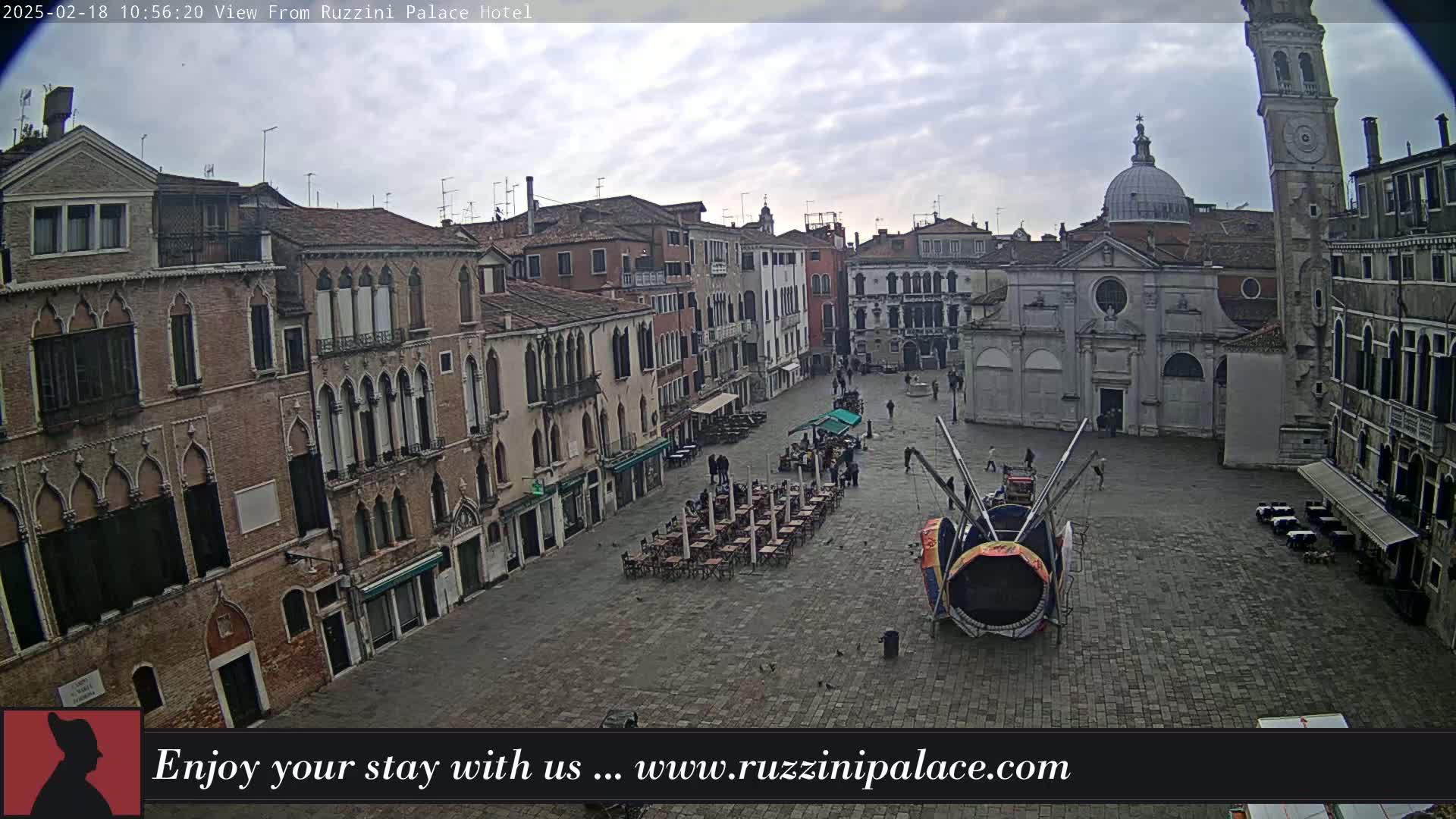 A large, paved square in Venice, Italy, is mostly empty under an overcast sky, with several buildings surrounding it, a few people, some outdoor seating, and a large colorful object in the center.