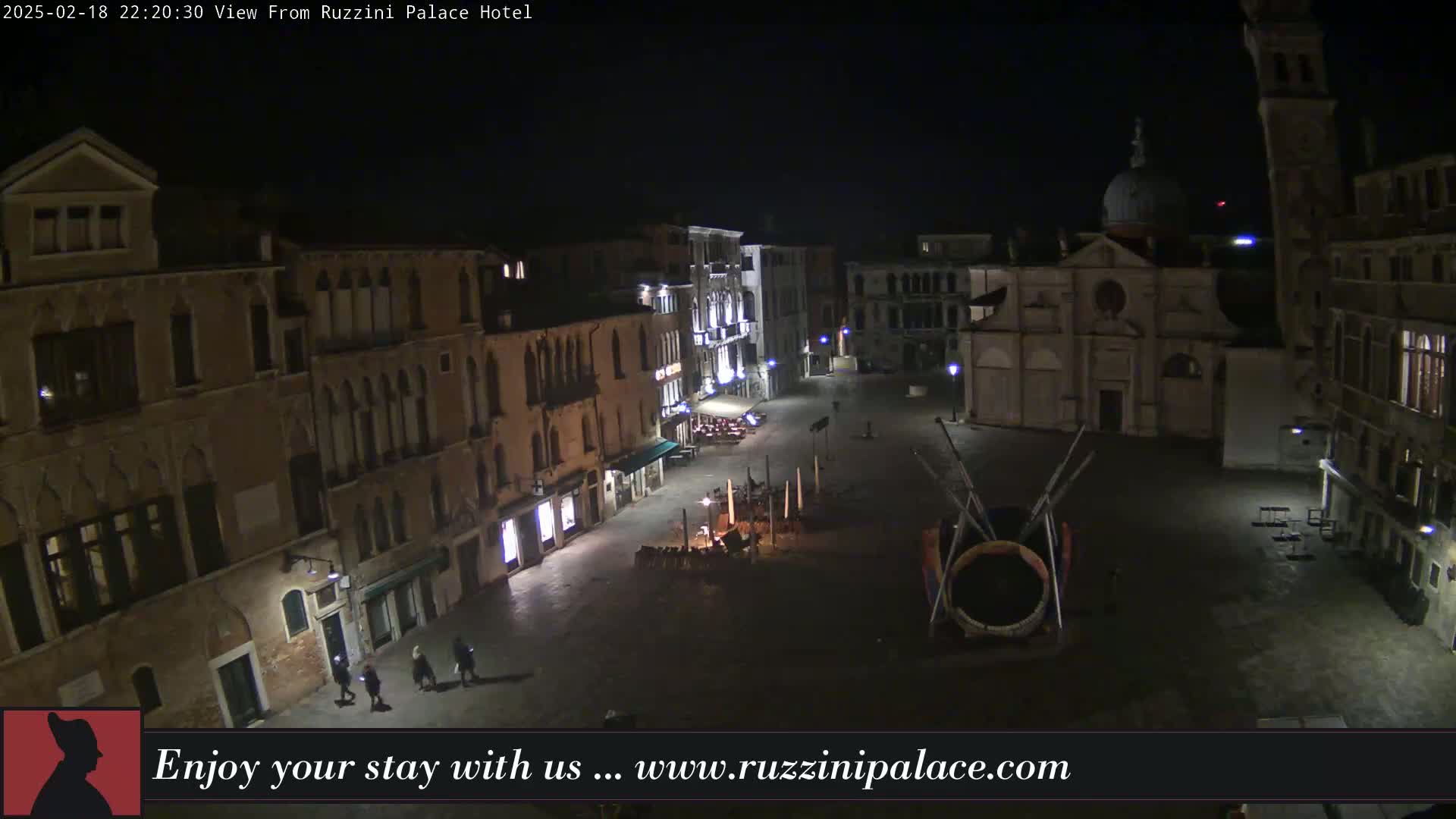 A nighttime, high-angle view shows a dimly lit, empty square in Venice, Italy, surrounded by old buildings, with a few pedestrians visible.