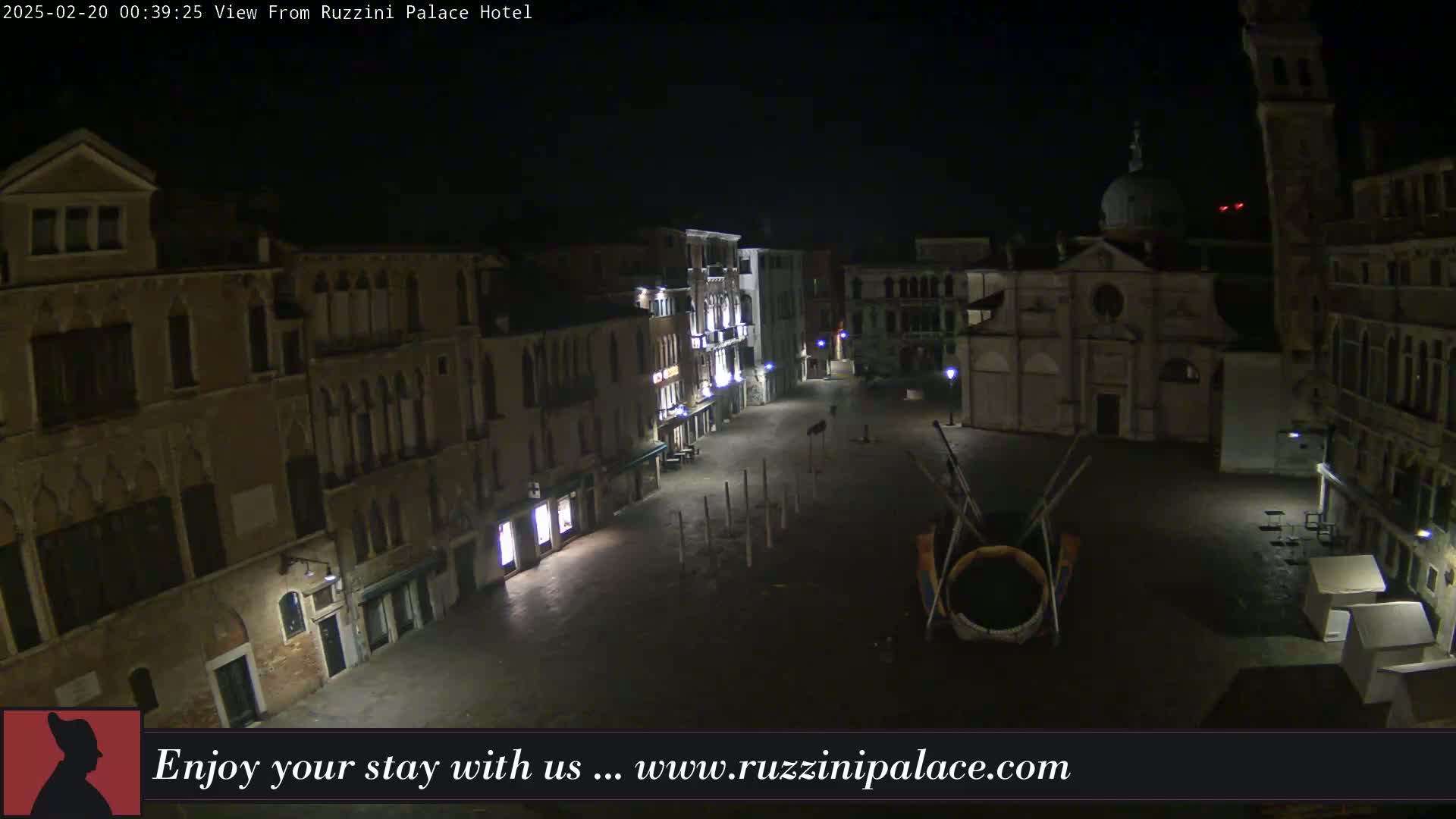 A nighttime, outdoor view of a dimly lit, empty square in Venice, surrounded by old buildings, featuring a large circular art installation in the center.