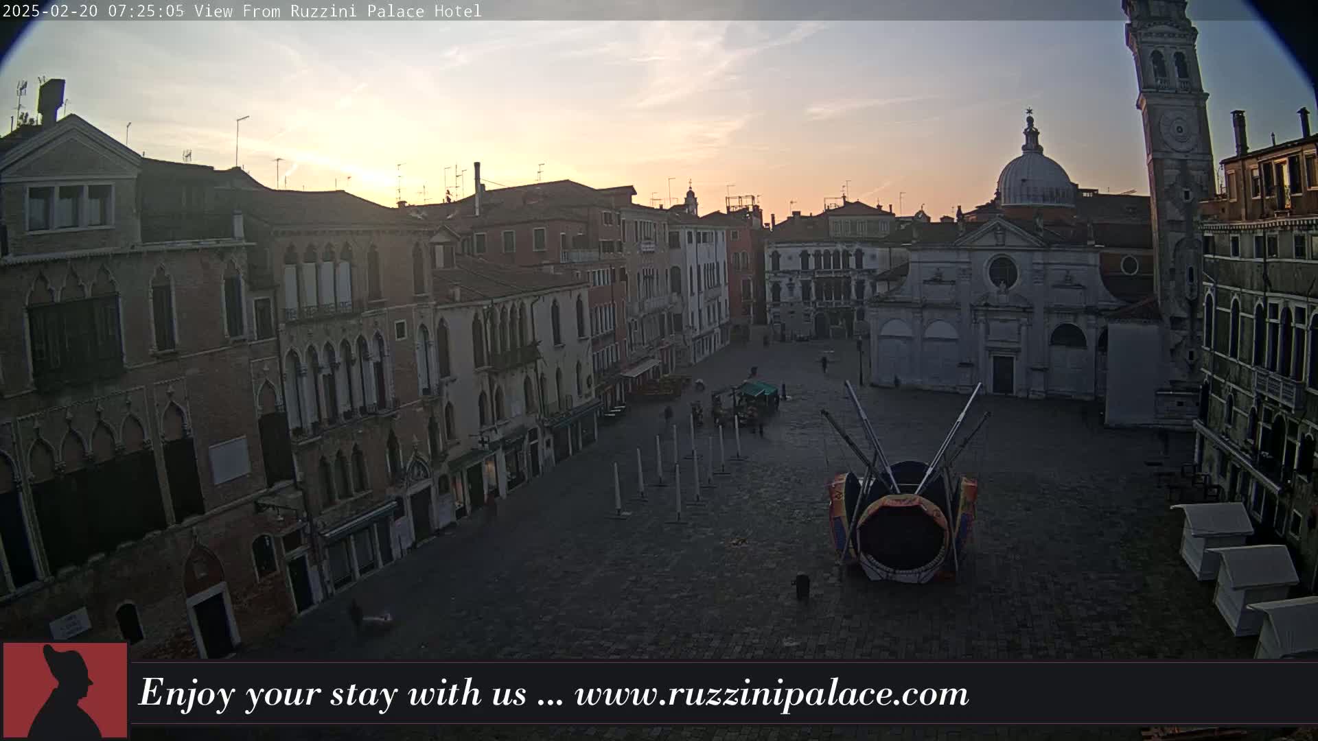 A sunrise illuminates a large, empty square in Venice, surrounded by aged brick buildings, with a carnival ride in the center.