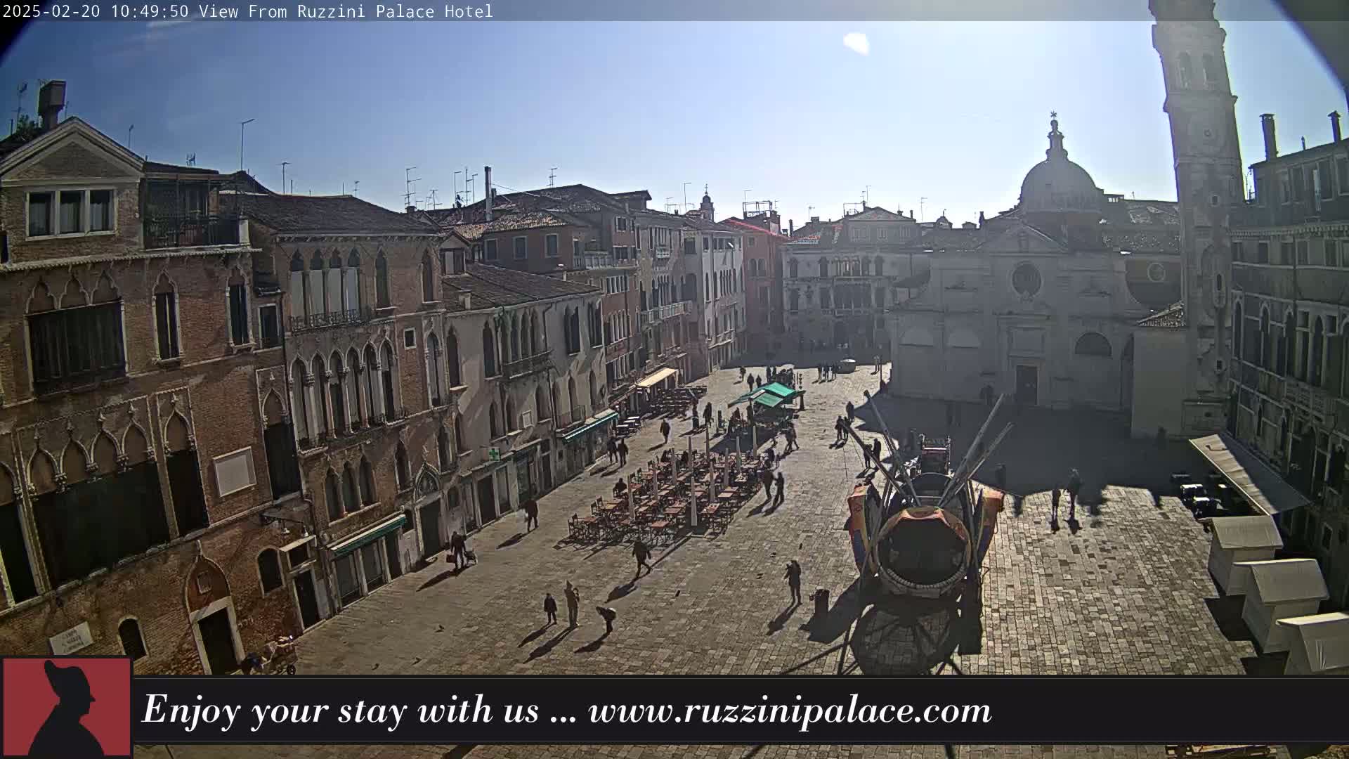 A sunny day reveals a large, paved square in Venice, Italy, surrounded by historic buildings, with some people walking and outdoor seating areas visible.