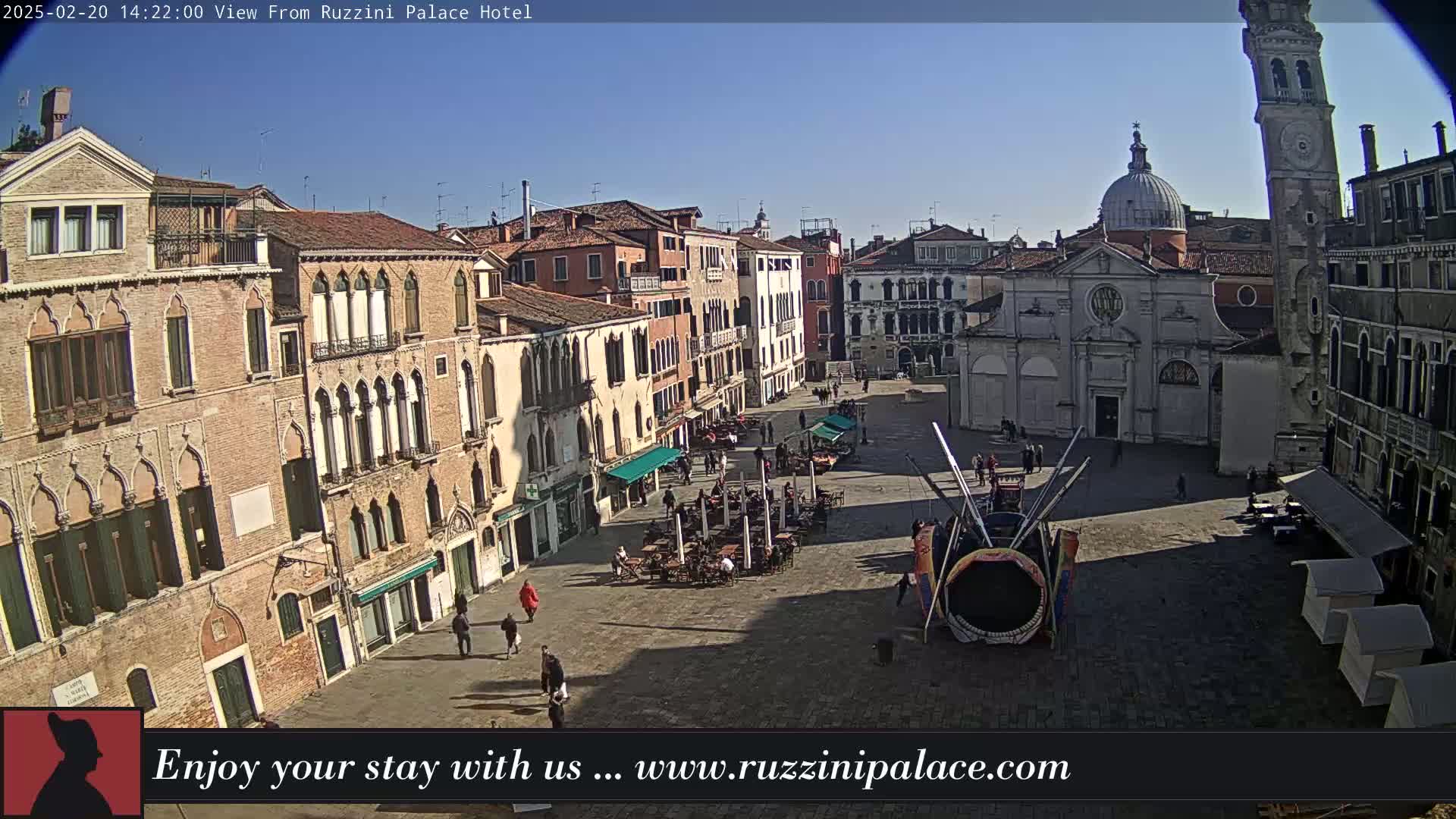 A sunny Venetian square is viewed from above, showing several buildings, a church, some outdoor seating areas, and a large carnival-type structure in the center.