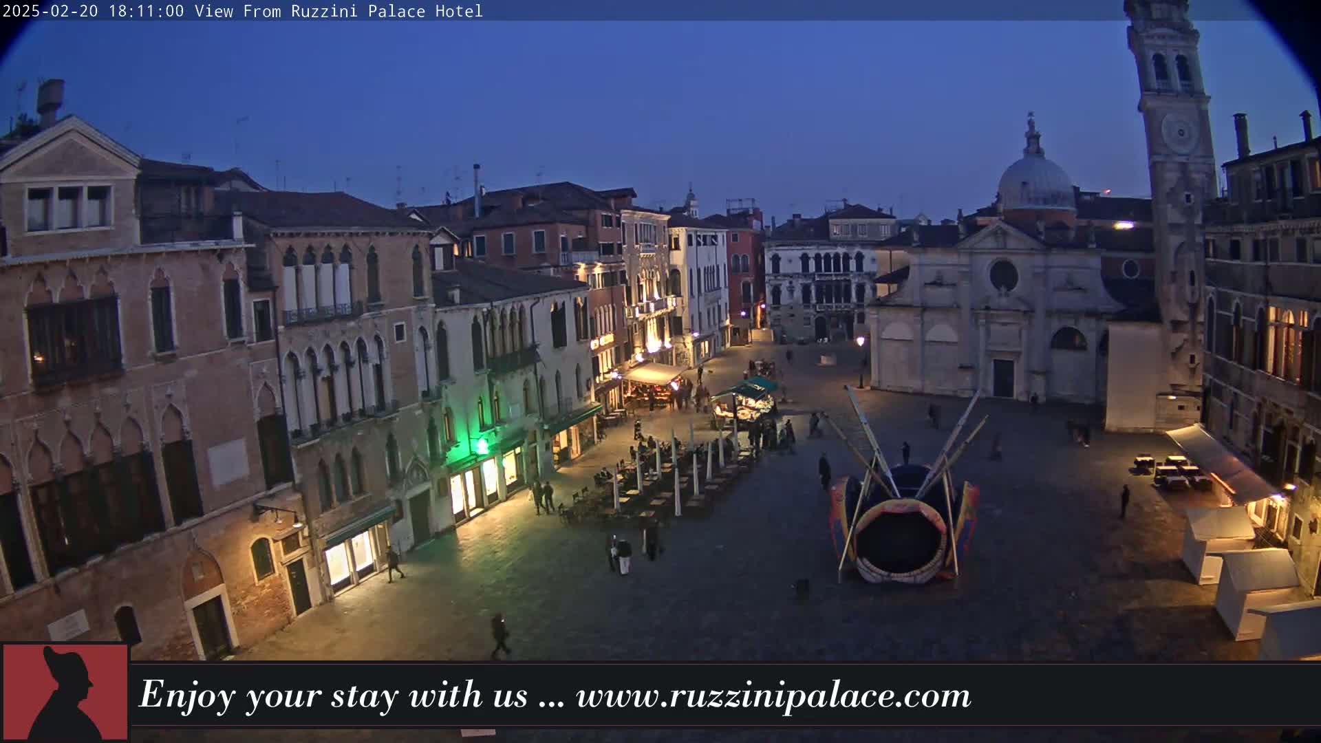 A twilight view of a cobblestone square in Venice, Italy, surrounded by old buildings, with a few people and outdoor seating visible.