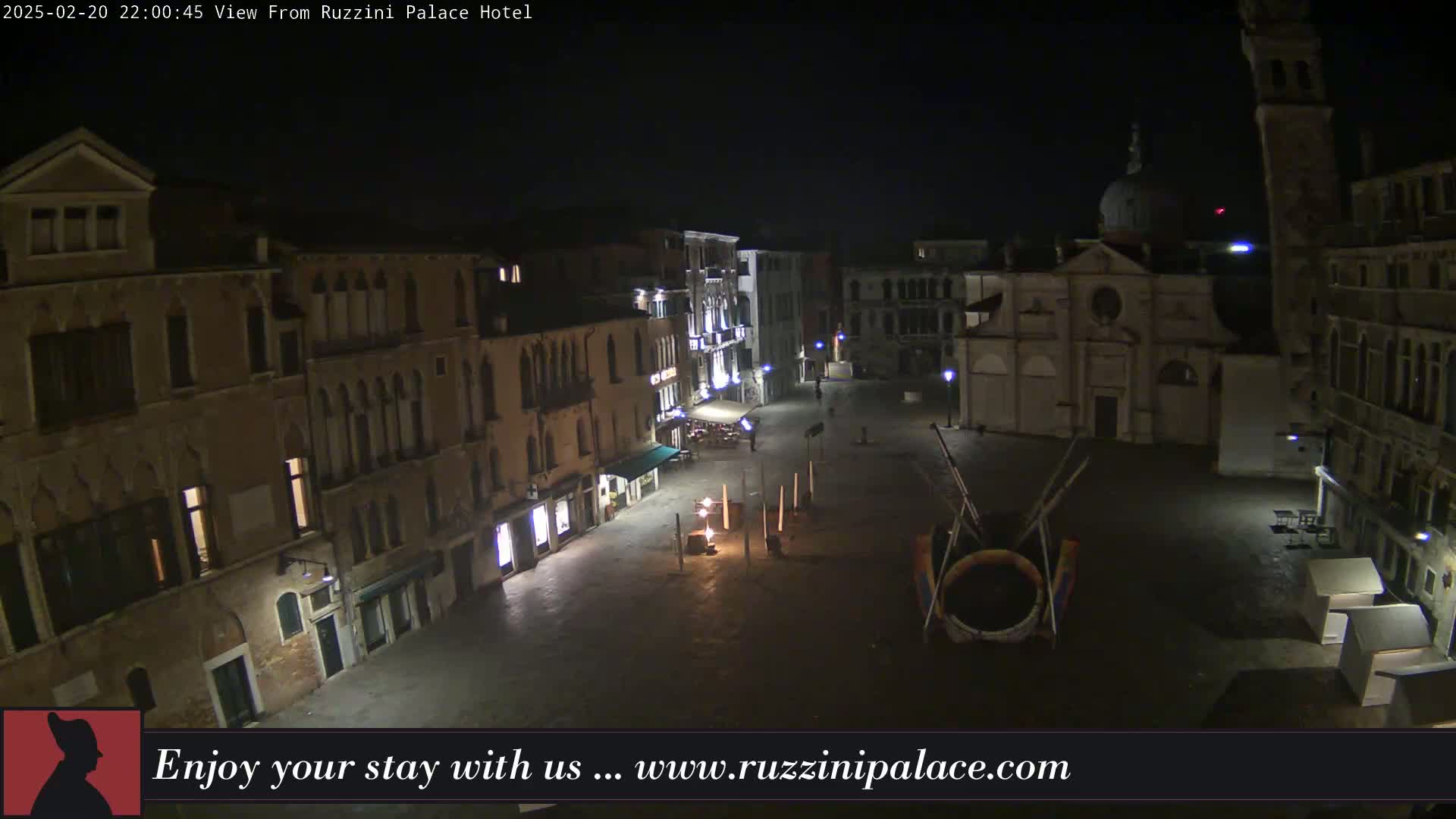 A nighttime, aerial view of a mostly empty, cobblestone plaza surrounded by old, multi-story buildings.