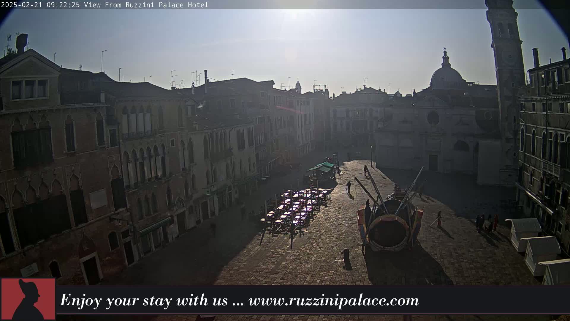 A sunny, outdoor view of a large plaza in Venice, Italy, shows several buildings surrounding it, with some people and outdoor seating visible.