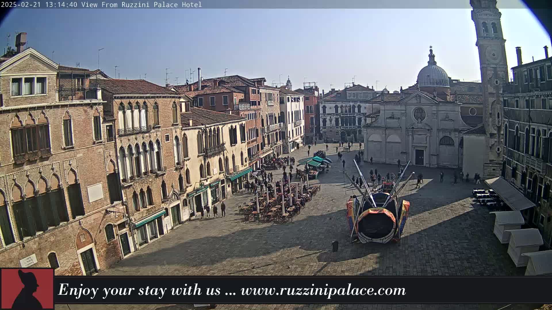 A sunny Venetian square is viewed from above, showing several buildings, a large outdoor seating area, and a colorful, large art installation in the center.