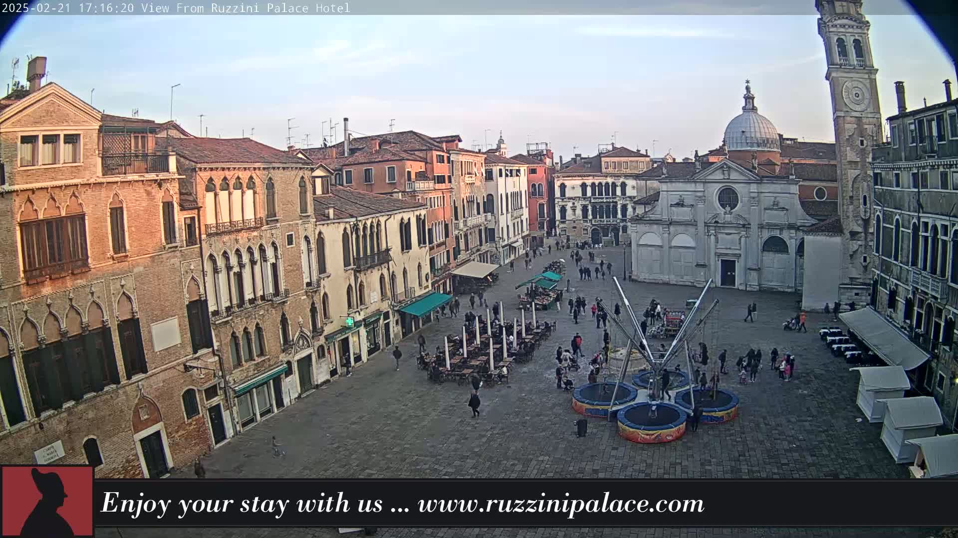 A mostly clear, twilight view of a large, cobblestone square in Venice, Italy, surrounded by historic buildings, with several people and outdoor seating areas.