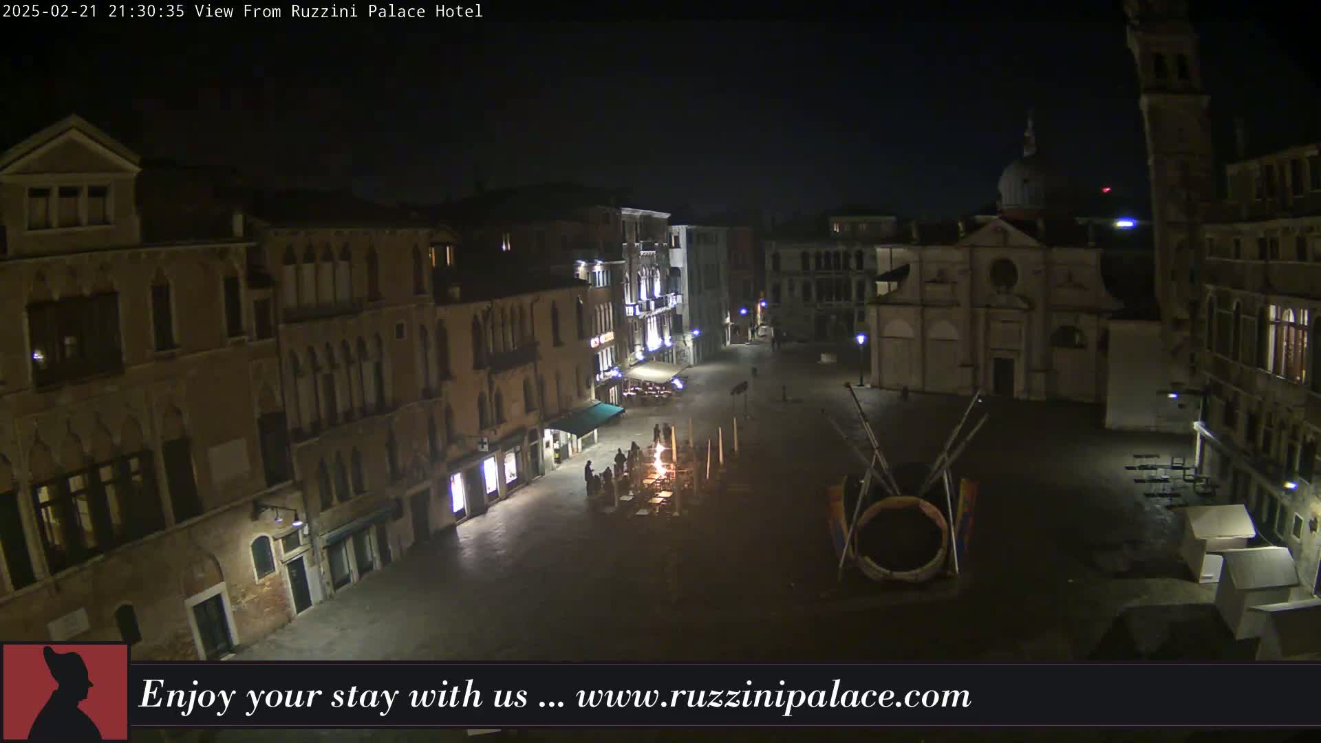 A nighttime, high-angle view of a dimly lit plaza in Venice, Italy, surrounded by old buildings, with a small gathering of people near a central light source.