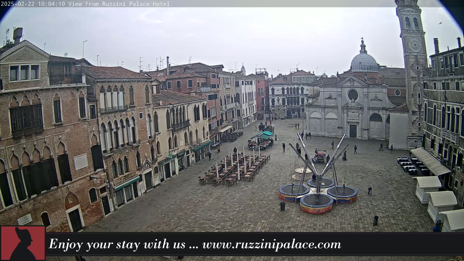 A large, paved square in a city is populated by a few people, several outdoor cafes, and some inflatable trampolines under an overcast sky.