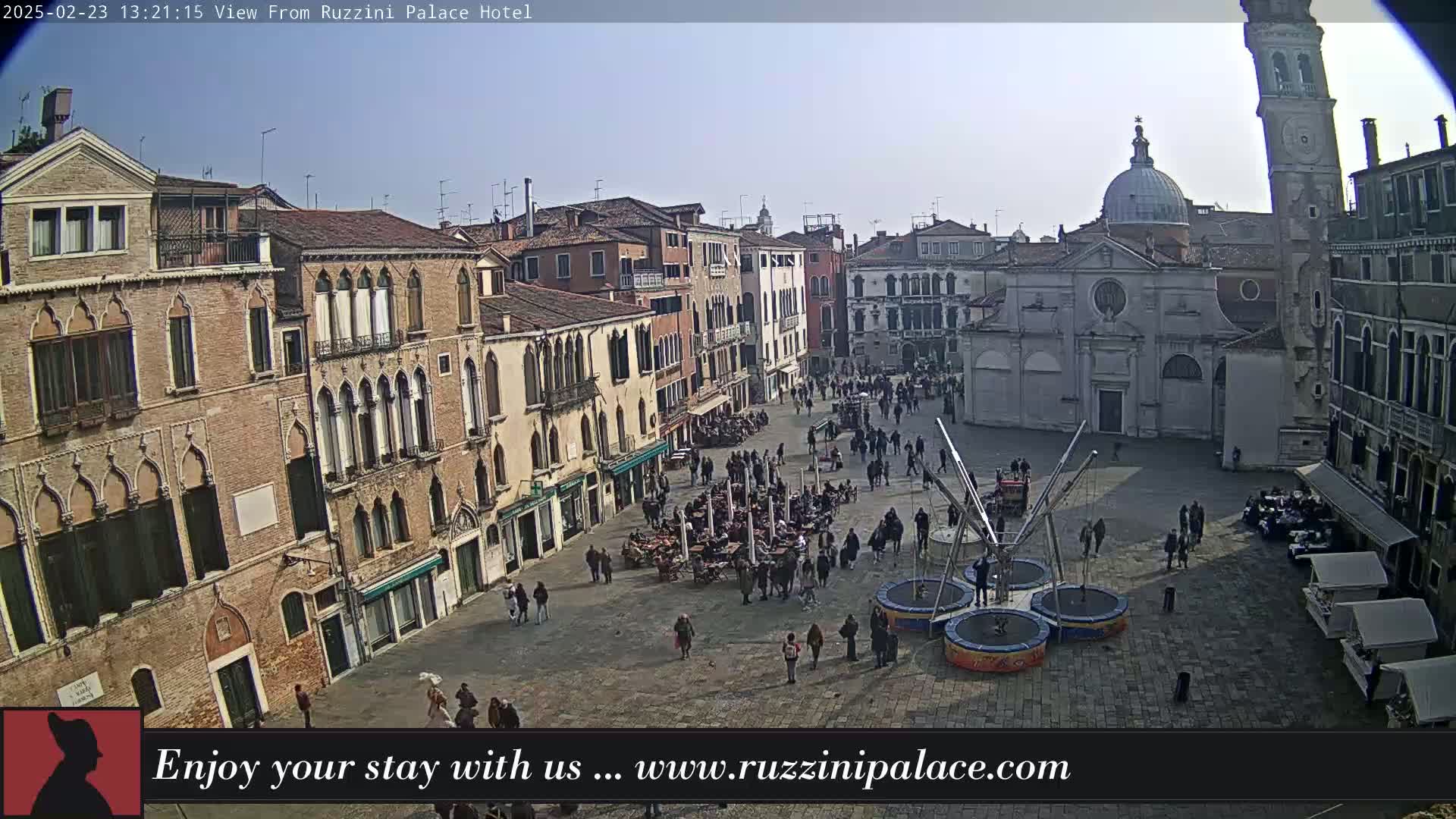 A sunny Venetian square is populated with people, outdoor seating, and a small amusement ride.