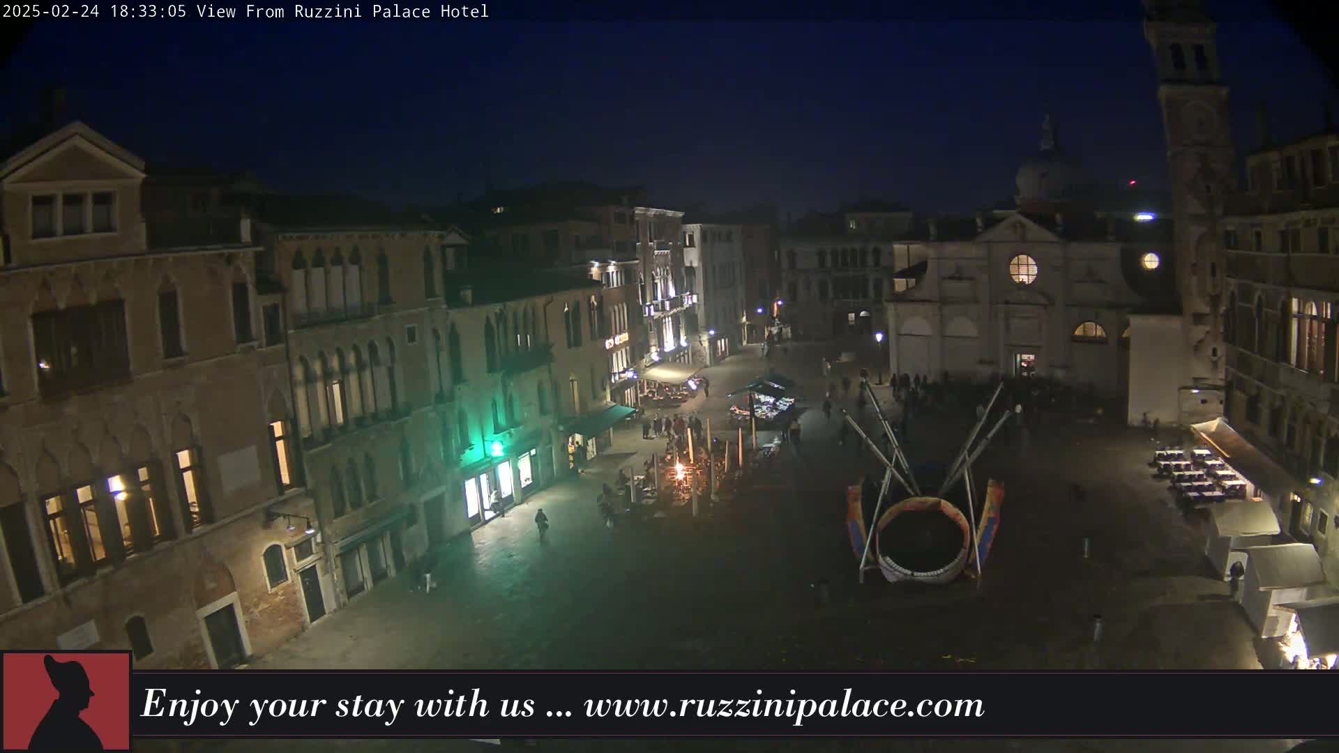 A nighttime aerial view of a European-style square with several buildings, some people, and outdoor seating areas under a dark sky.