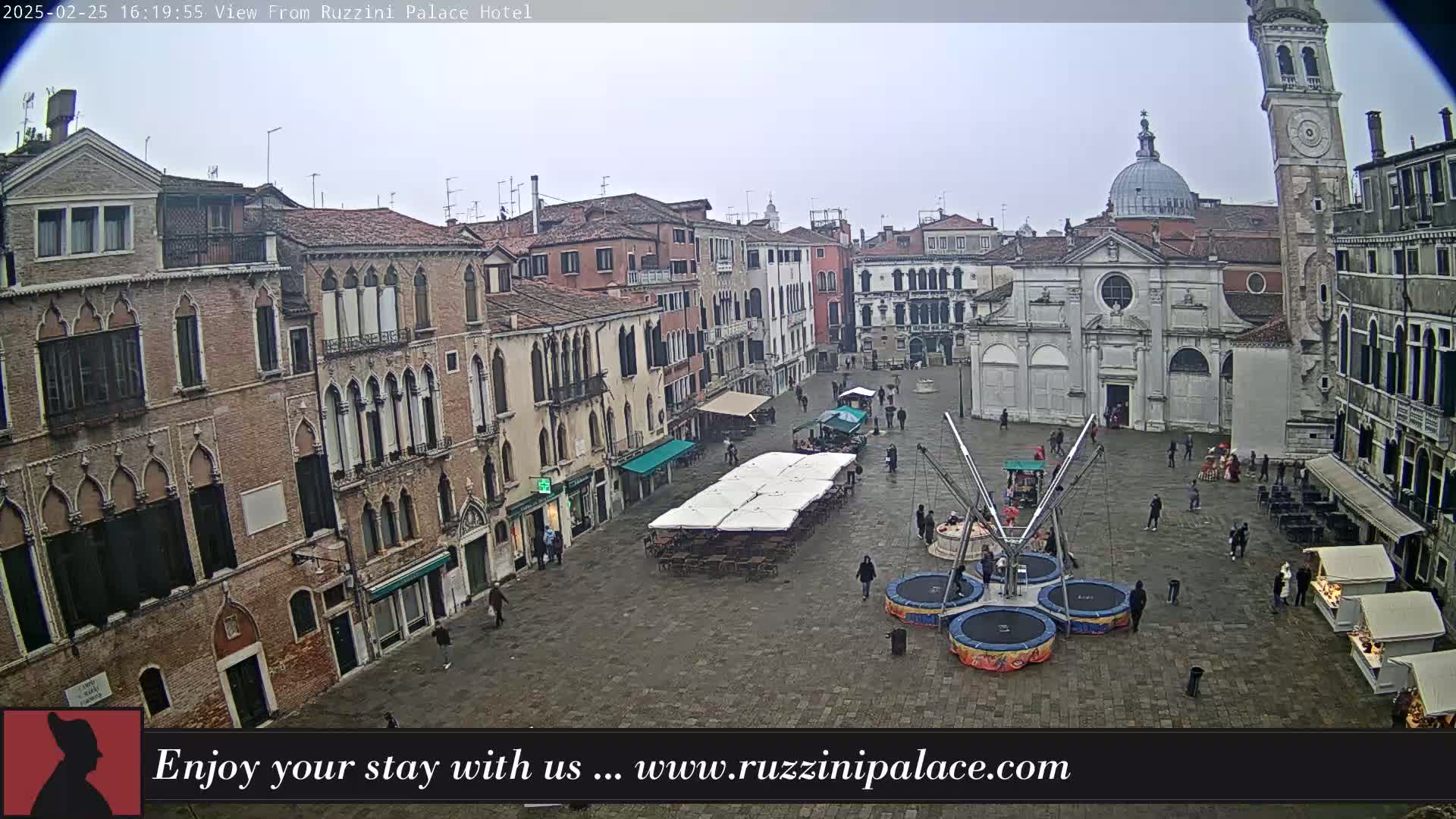 A large, stone plaza in a city is filled with several small temporary structures, a few people walking around, and a small amusement park ride; the overcast sky suggests a cloudy day.