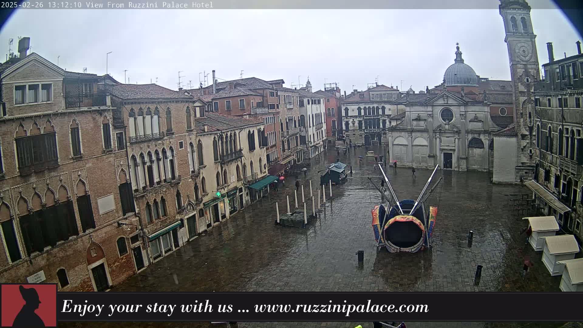 A wet, stone plaza in Venice, Italy, is surrounded by tall buildings, one of which has a dome, and a carnival ride sits in the center.