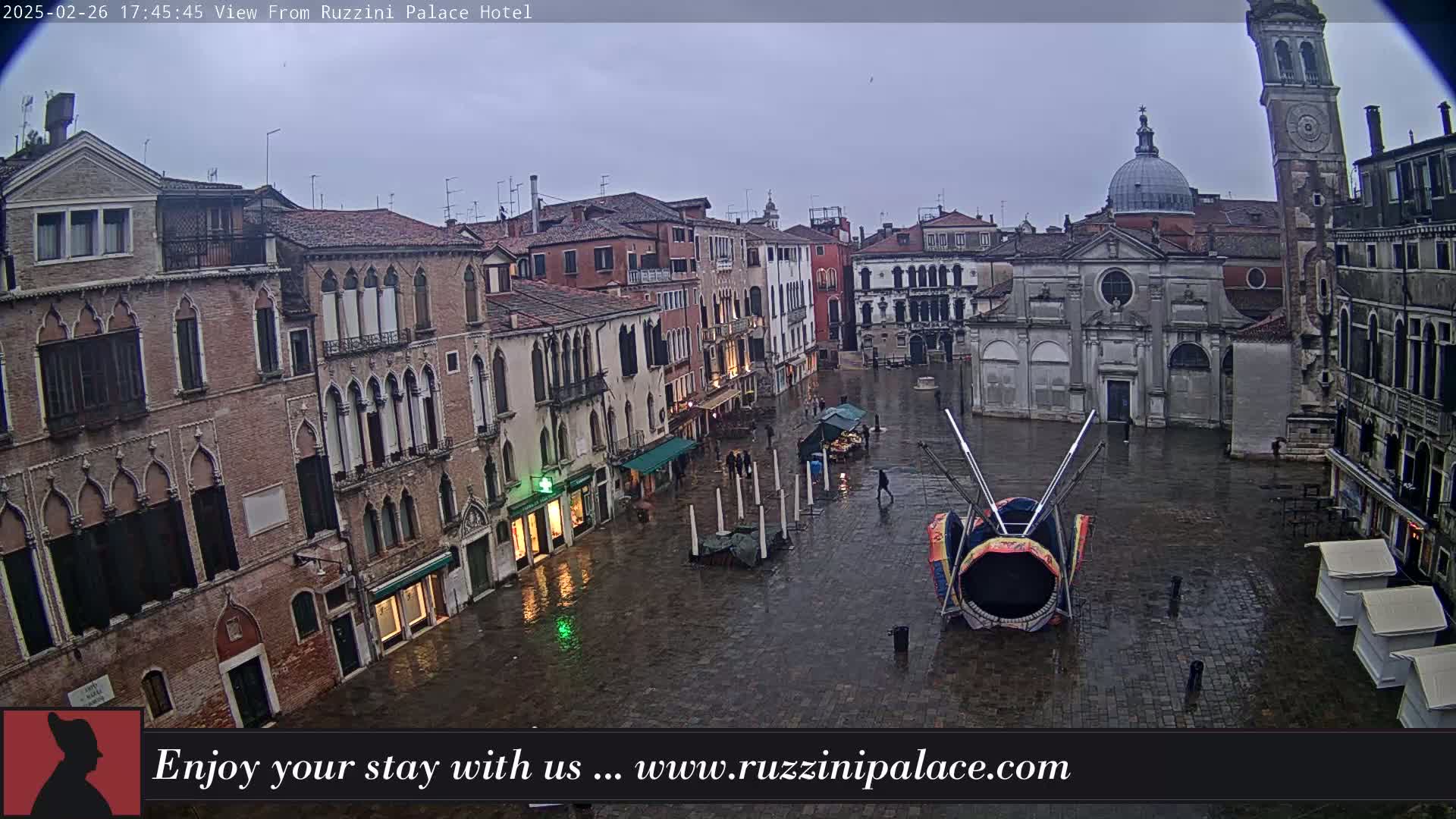 A wet, stone plaza in Venice, Italy, is surrounded by old buildings, with a colorful, insect-like structure in the center.