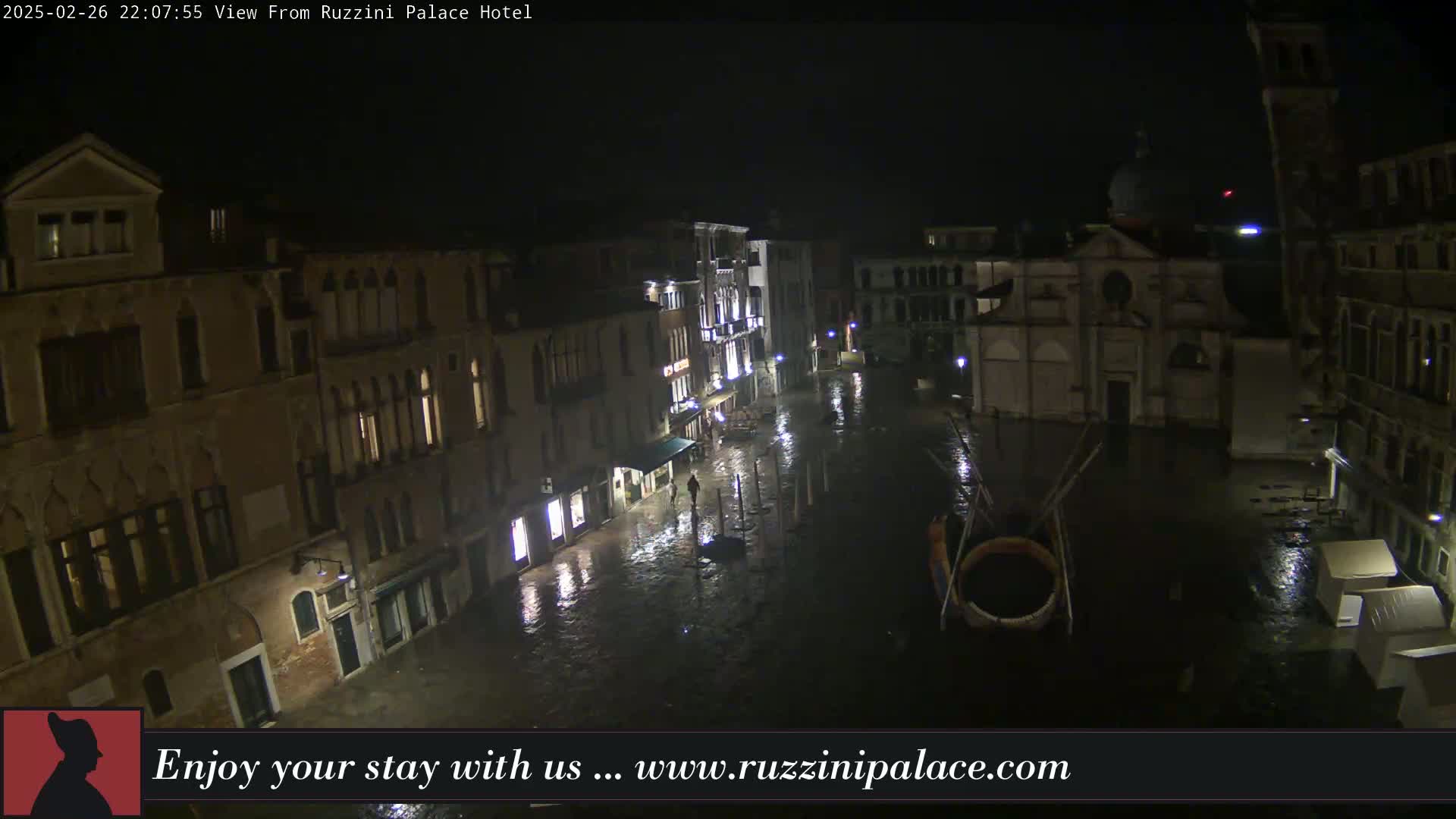 A nighttime view of a rain-soaked Venetian square, showing several buildings with illuminated windows and a church in the background.