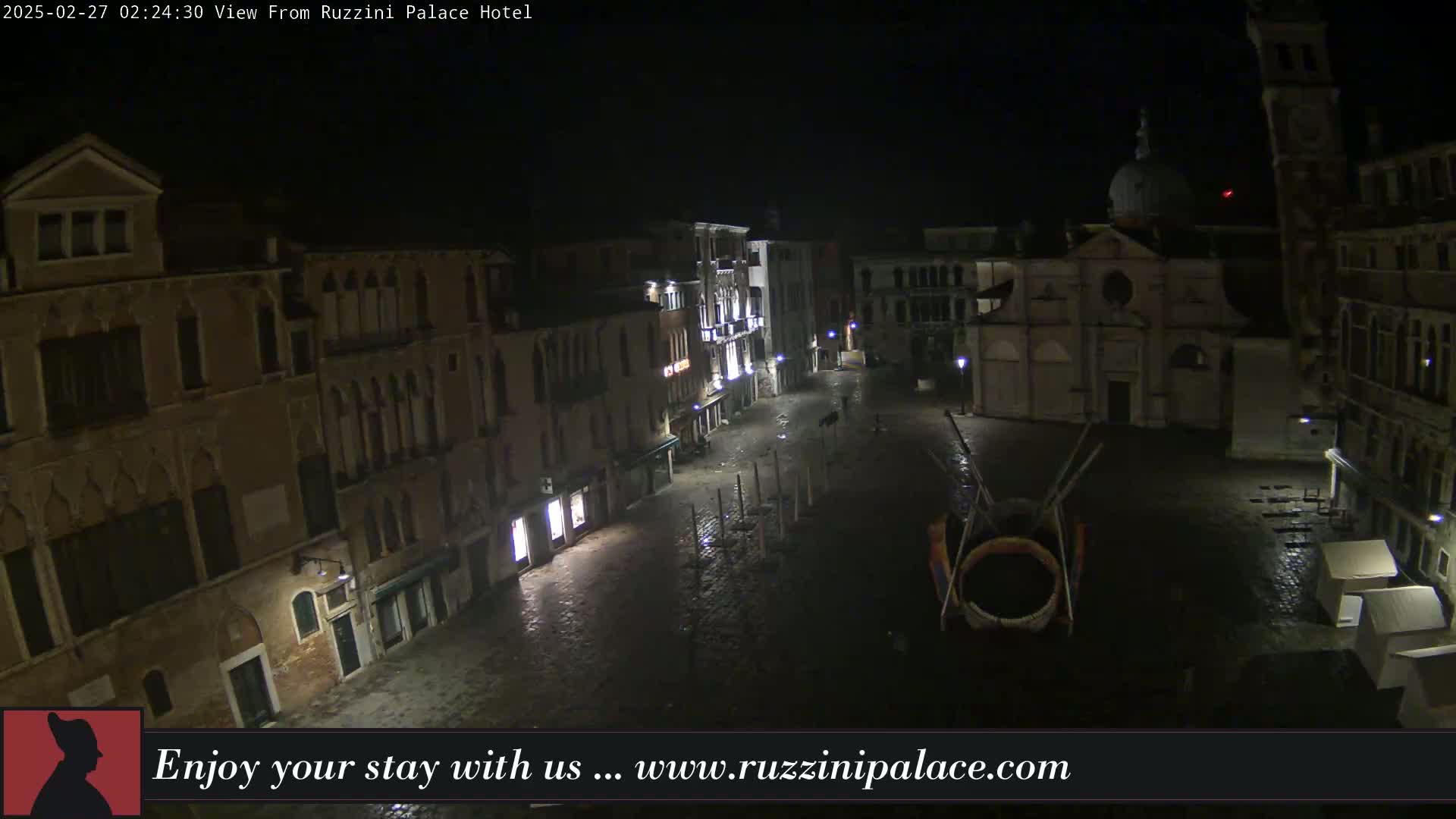 A nighttime, wet, aerial view of a dimly lit European square surrounded by old buildings, featuring a large sculpture in the center.