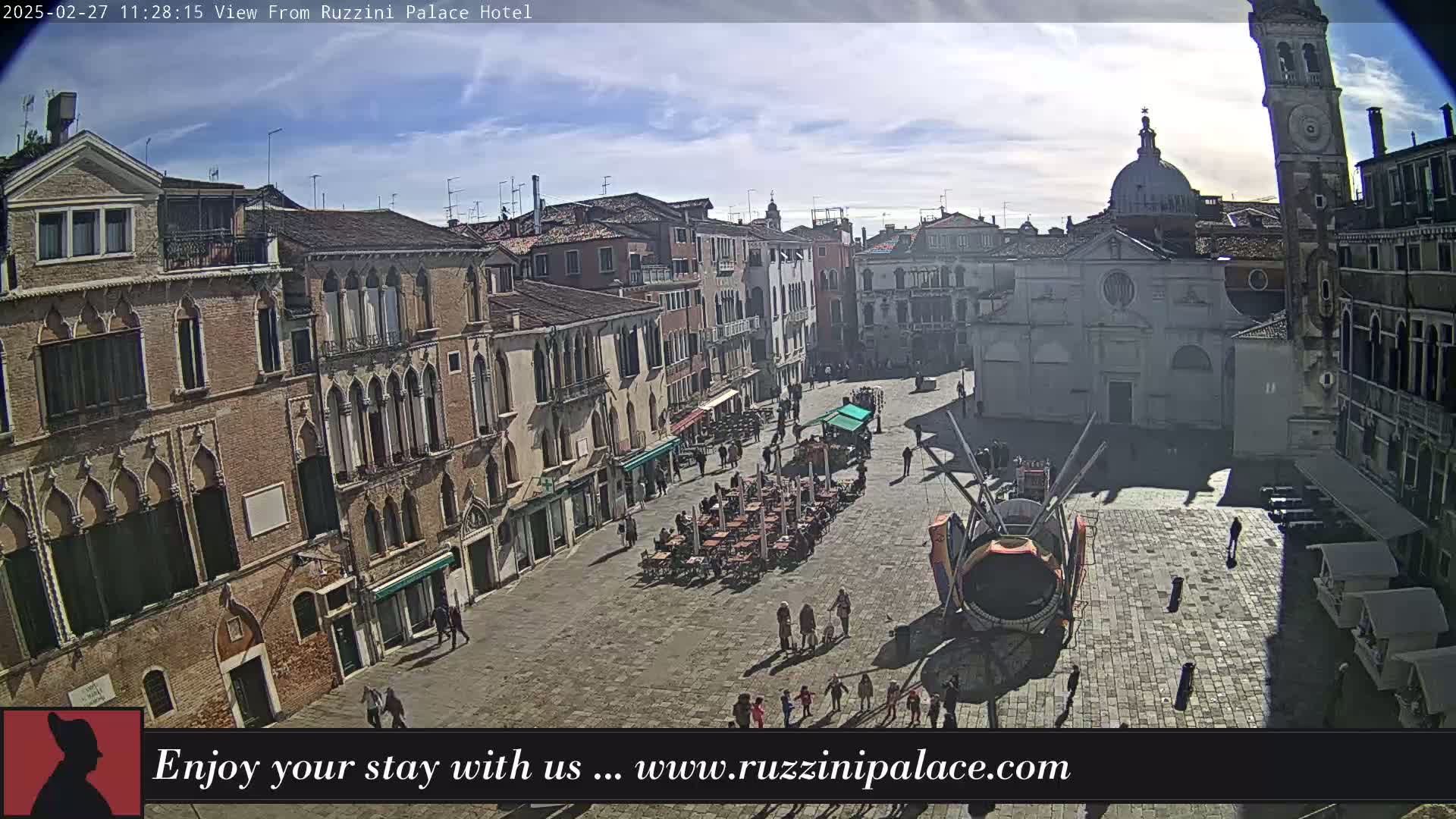 A sunny day reveals a large, paved square in Venice, surrounded by historic buildings, with several people and outdoor seating areas visible.