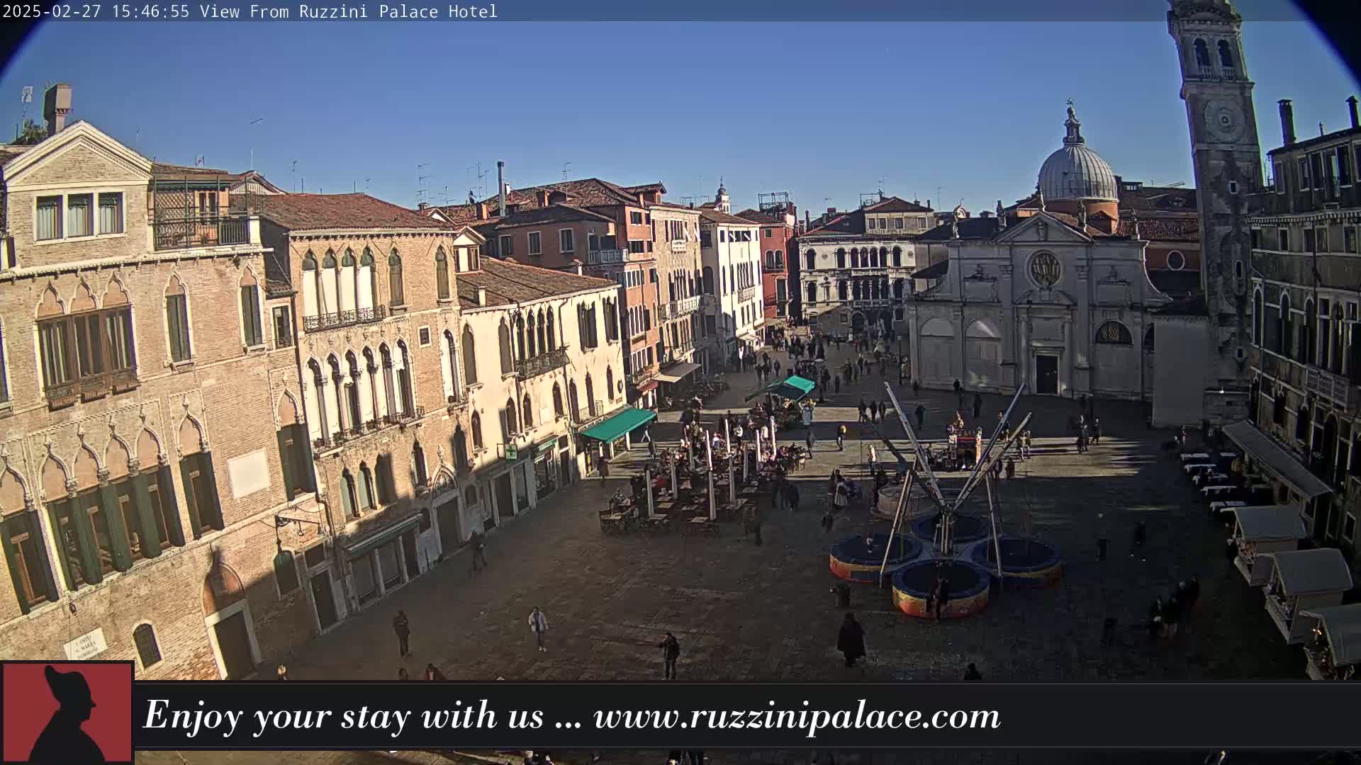 A sunny, outdoor view of a large town square in Venice, Italy, filled with people and featuring historic buildings, including a church with a dome and bell tower.