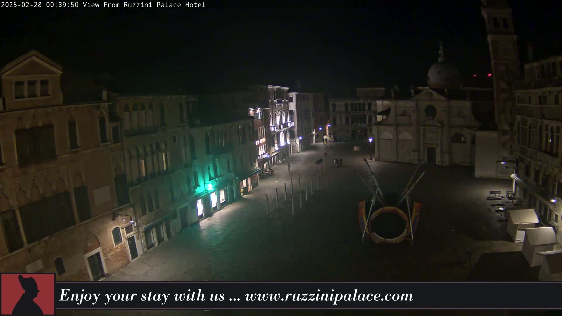 A nighttime view of a dimly lit Venetian square, featuring several buildings, a church, and sparse pedestrian activity under clear skies.