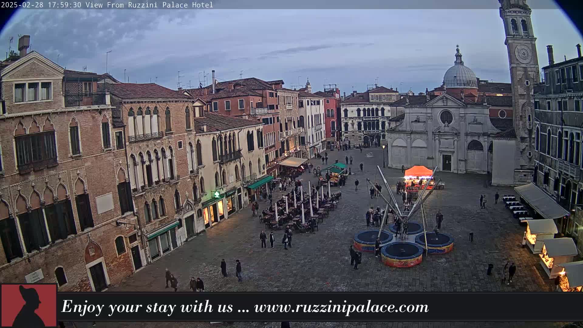 A twilight view of a large, cobblestone square in Venice, Italy, filled with people and outdoor seating, featuring a church, several buildings, and a small amusement park attraction.