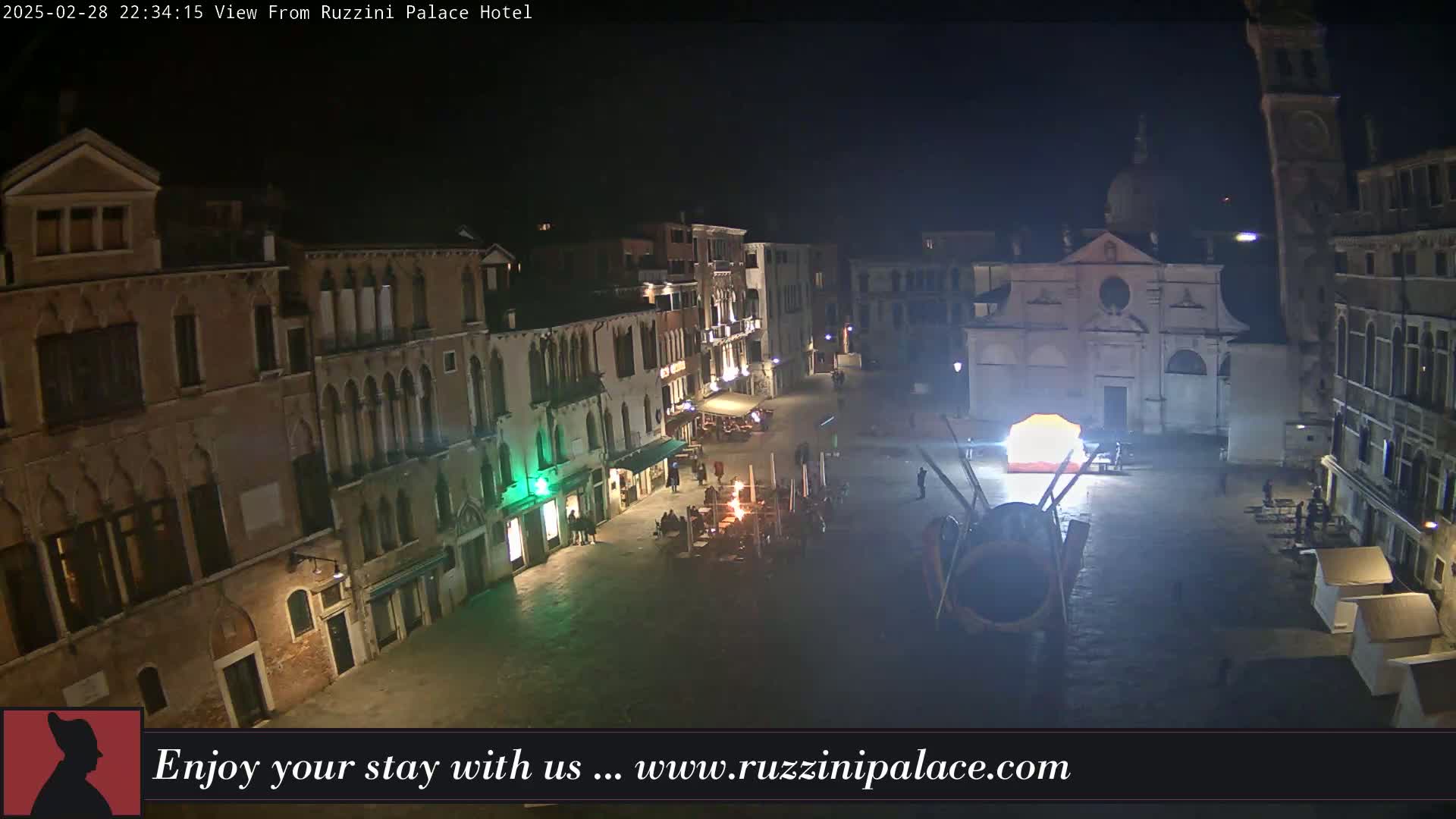 A nighttime, high-angle view of a dimly lit Venetian square with several buildings surrounding it, some with lights on, and what appears to be a temporary structure or installation in the center.