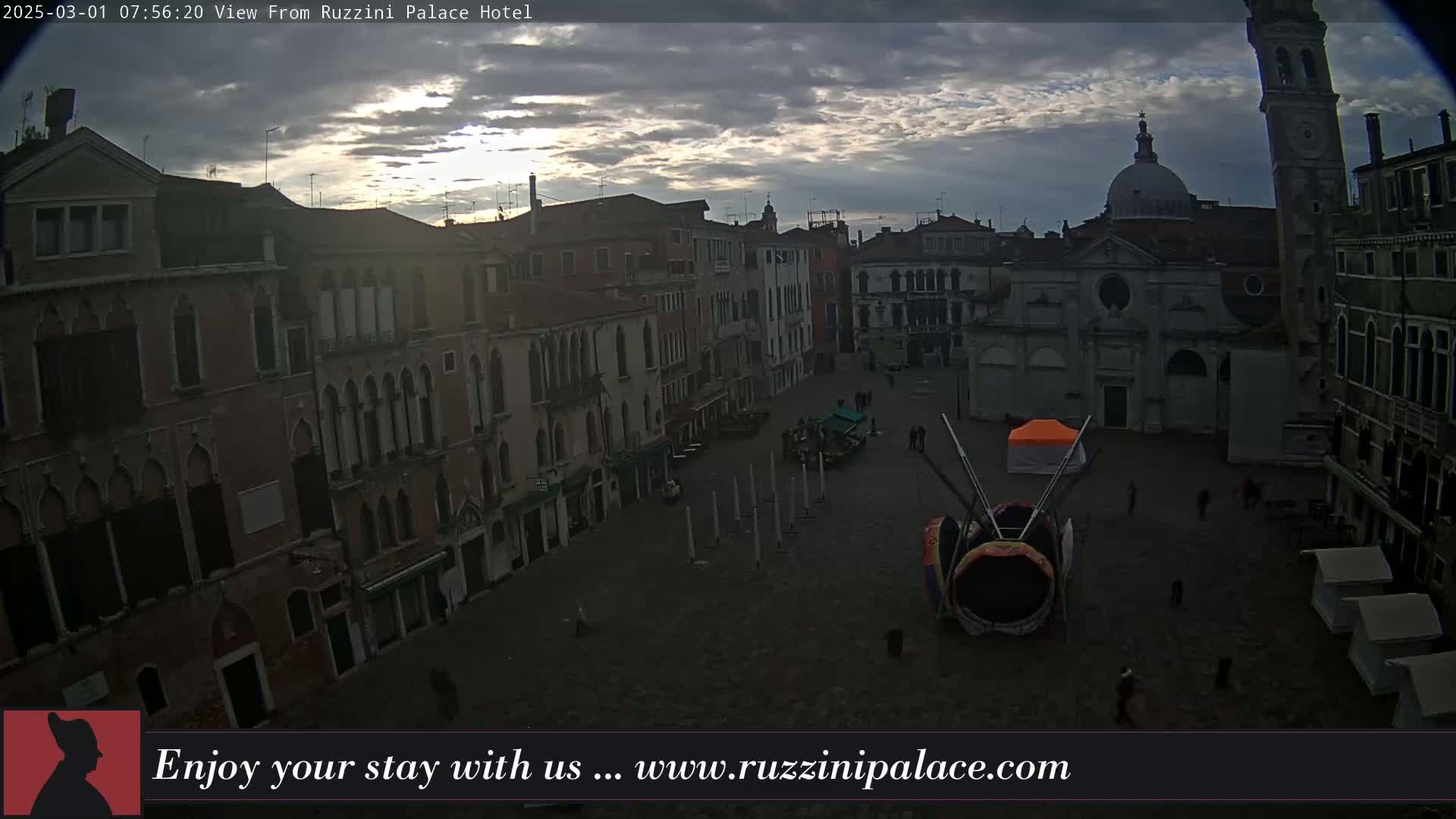 A mostly cloudy sunrise illuminates a large, empty square in Venice, Italy, surrounded by old buildings, with a large, colorful art installation in the center.