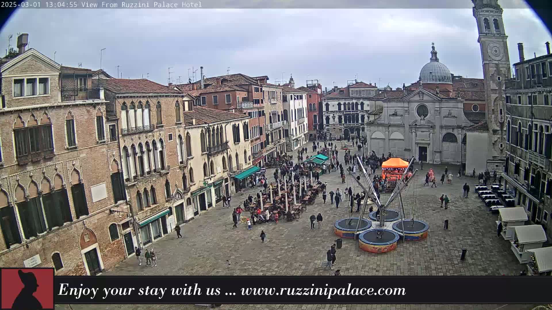 An overcast day reveals a large, paved square in Venice, Italy, surrounded by historic buildings, with several people walking around and some seated at outdoor cafes, and a small trampoline park in the center.