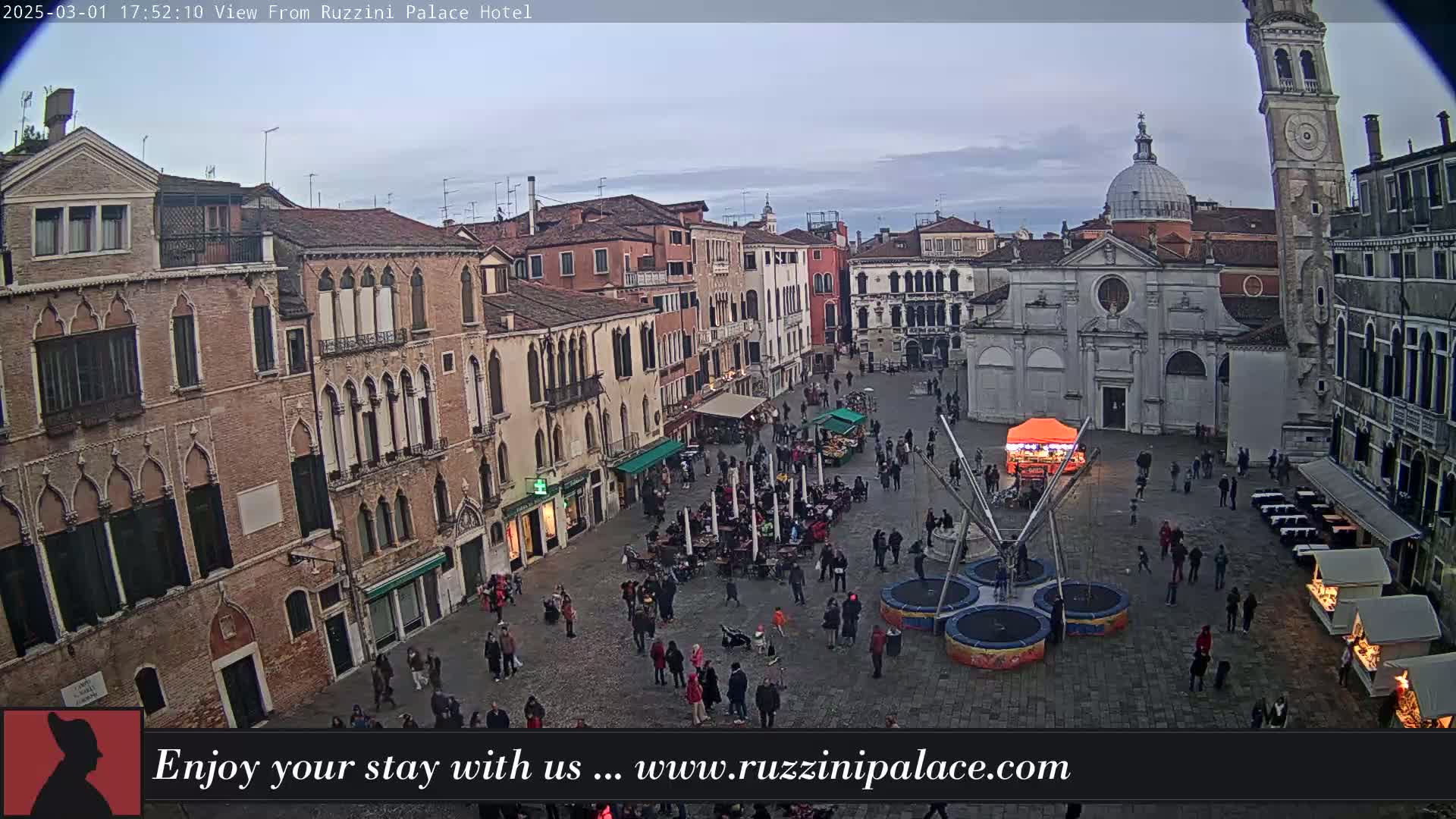 An overcast sky hangs over a large, stone plaza in Venice, Italy, where people are gathered around outdoor seating and a small amusement ride; several historic buildings surround the plaza.