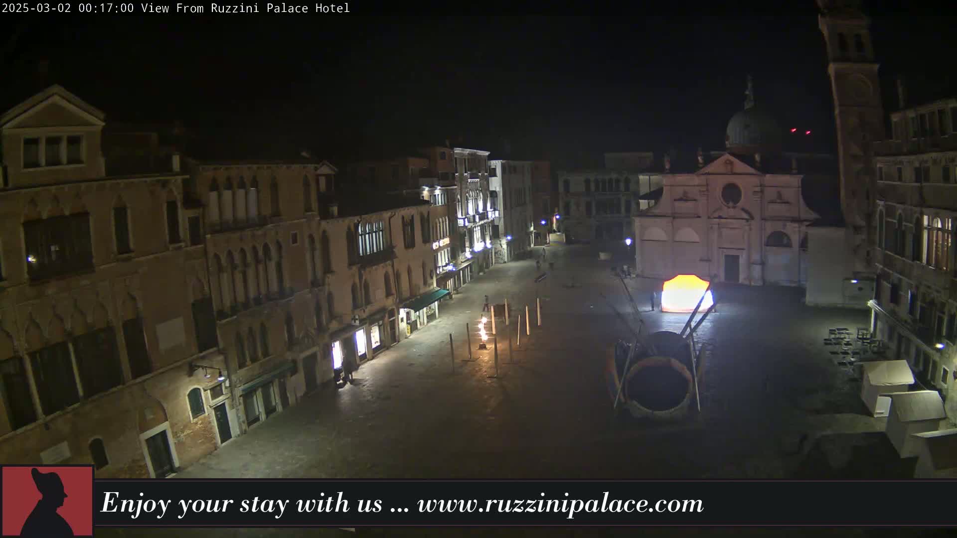 A nighttime view of a town square in Venice, Italy, shows several buildings illuminated with lights, a large art installation, and a mostly empty square.