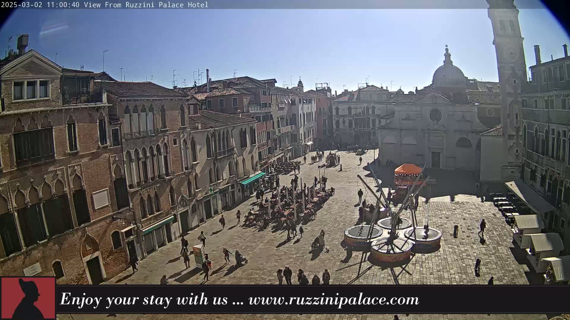 A sunny day reveals a large, paved square in Venice, surrounded by historic buildings, with people scattered throughout and amusement rides set up in the center.