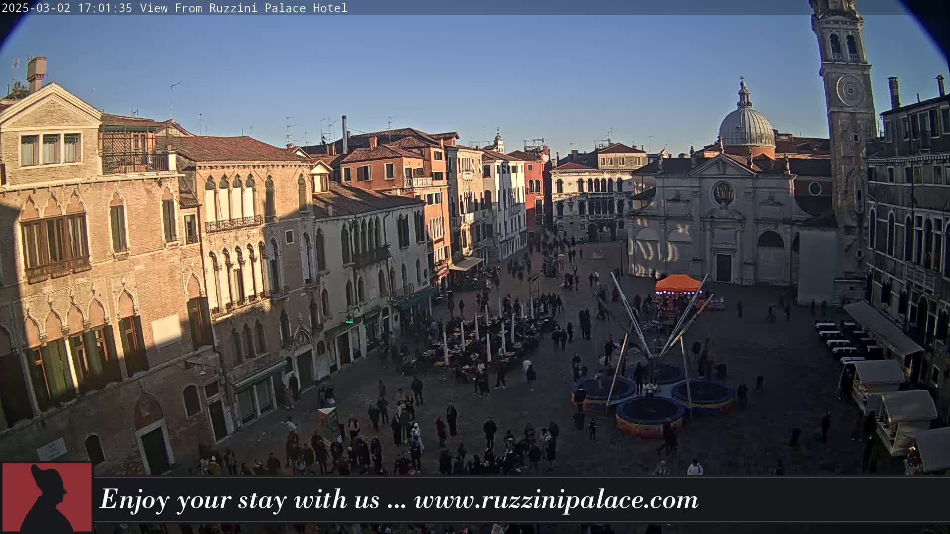 A sunny, clear day reveals a Venetian plaza filled with people and several buildings, including a church and a tall tower.