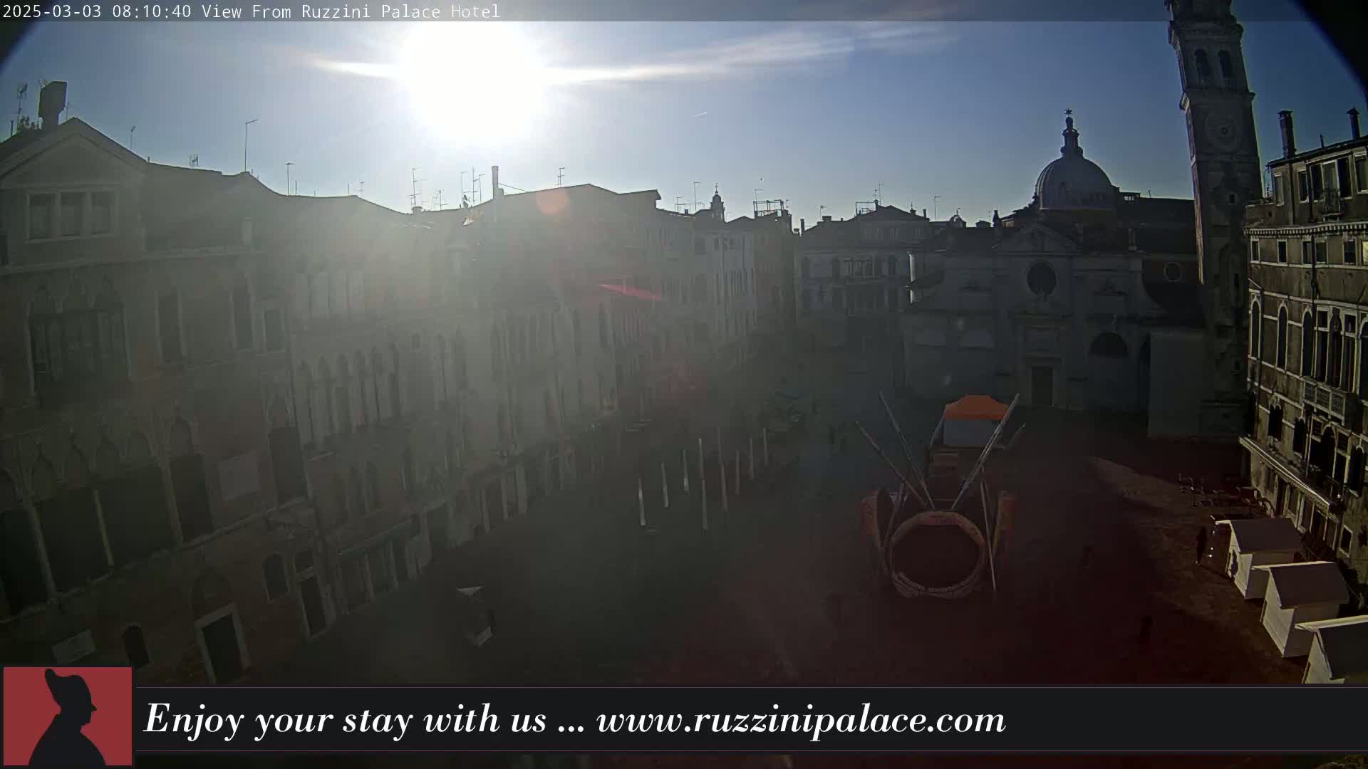 A sunny day reveals a Venetian square surrounded by old buildings, a tall tower, and a domed church, with some temporary structures in the plaza.