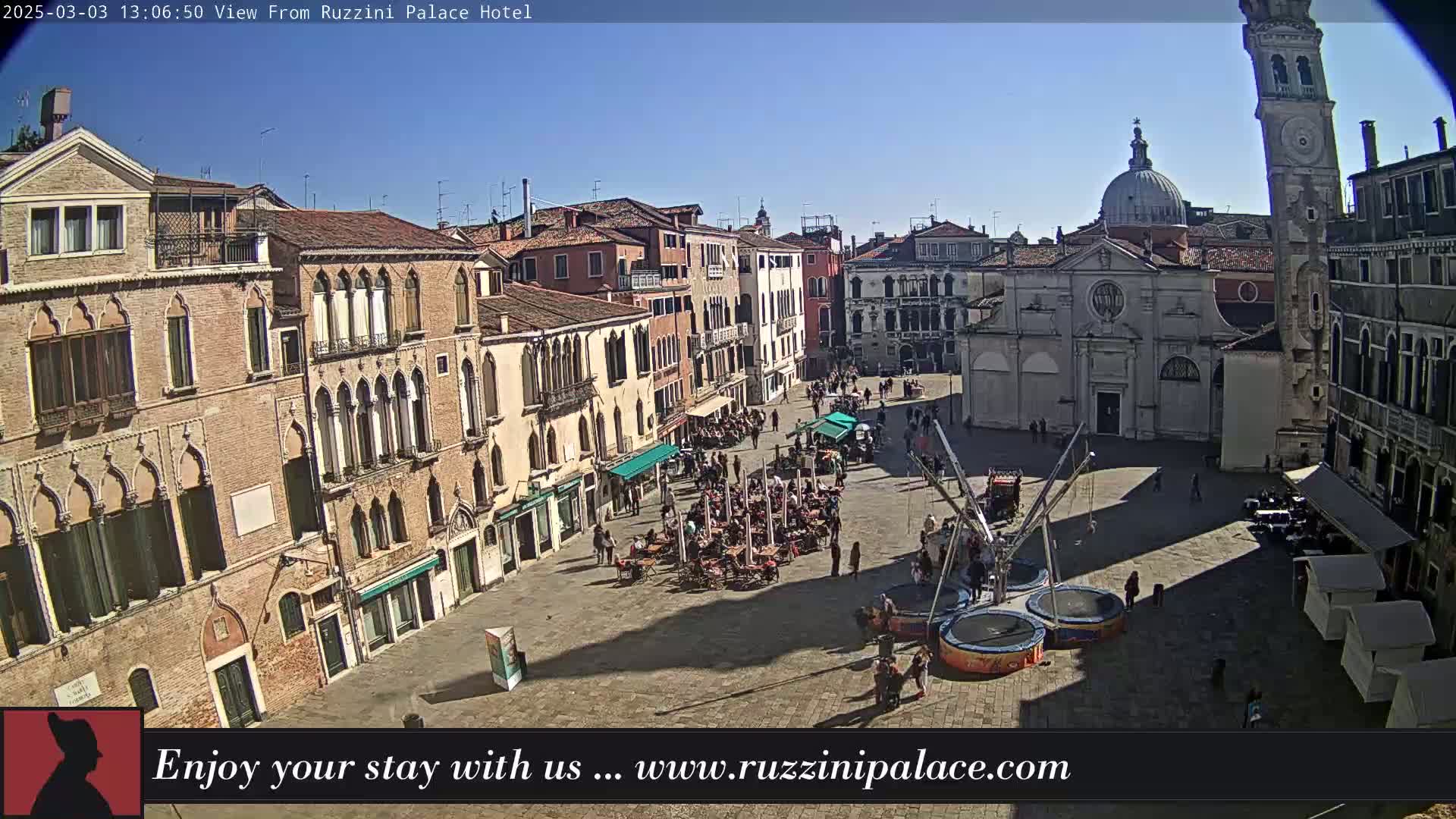 A sunny Venetian piazza is shown from an elevated viewpoint, with several buildings surrounding a paved square containing outdoor seating, a small bouncy-castle type attraction, and a scattering of people.