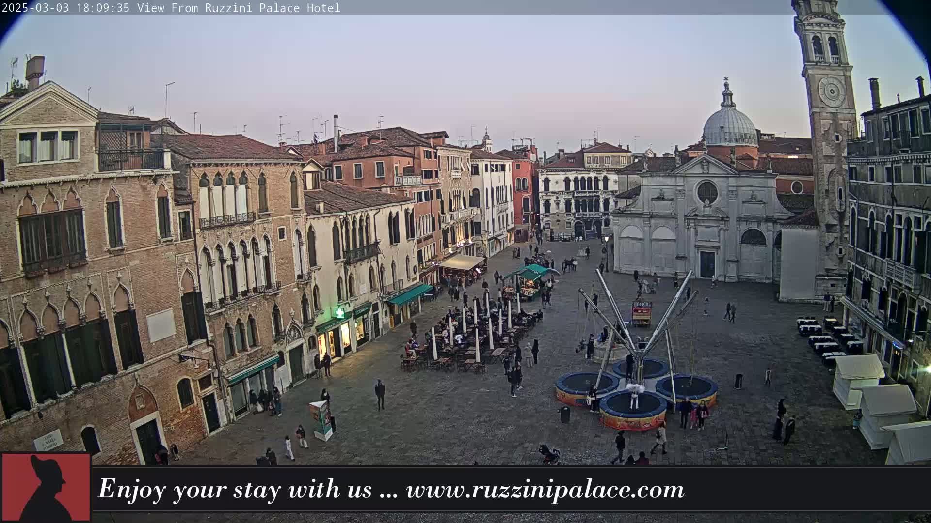 An outdoor, twilight view of a large, paved square in Venice, Italy, surrounded by old buildings, with several people and some outdoor seating.