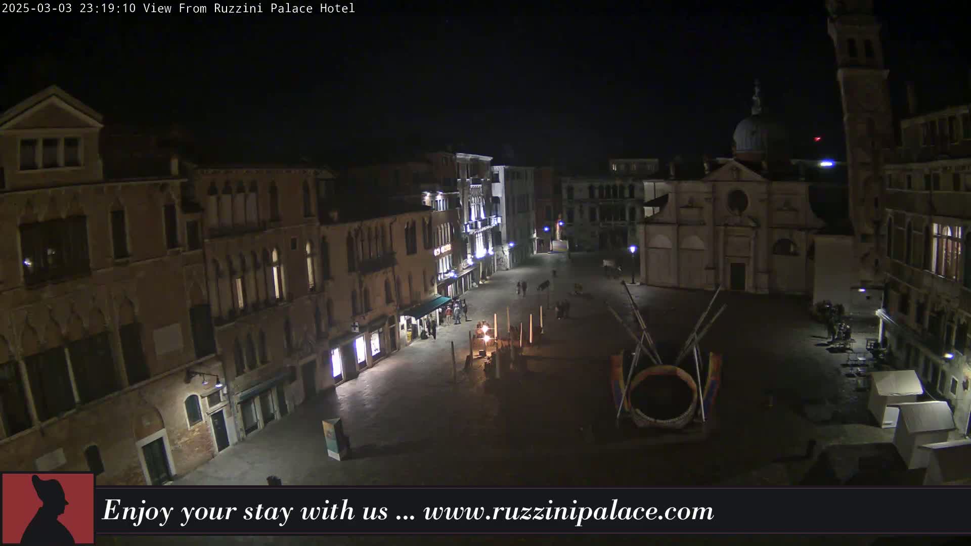 A nighttime aerial view of a dimly lit, mostly empty town square in Venice, featuring several buildings and a church.
