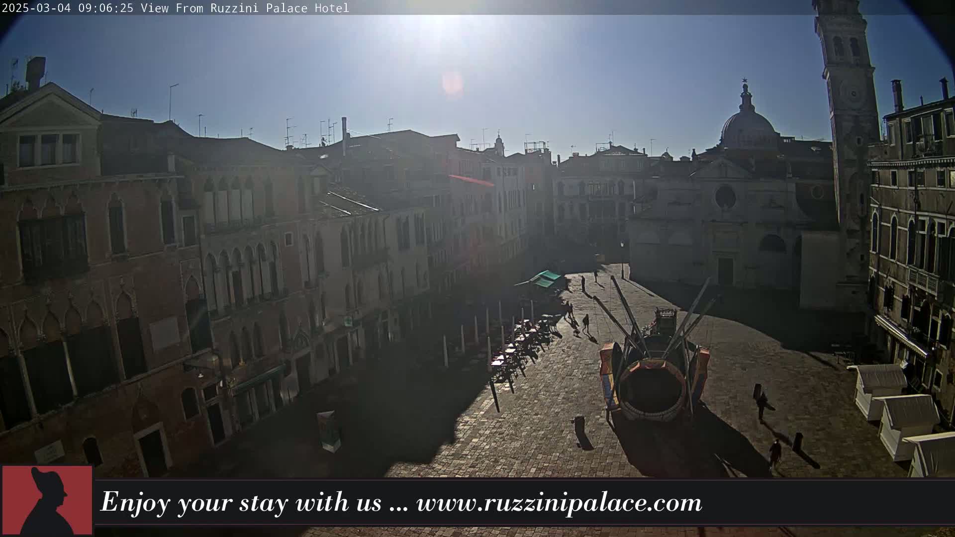 A sunny day reveals a Venetian square with several buildings, a large structure resembling an insect, and a few people sparsely scattered throughout.