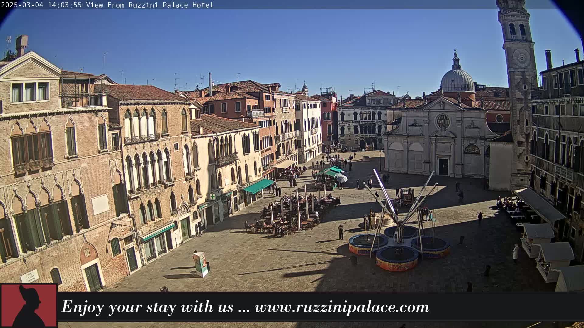 A sunny day reveals a large plaza in Venice, Italy, filled with people, outdoor seating, and a bouncy attraction, surrounded by historic buildings.