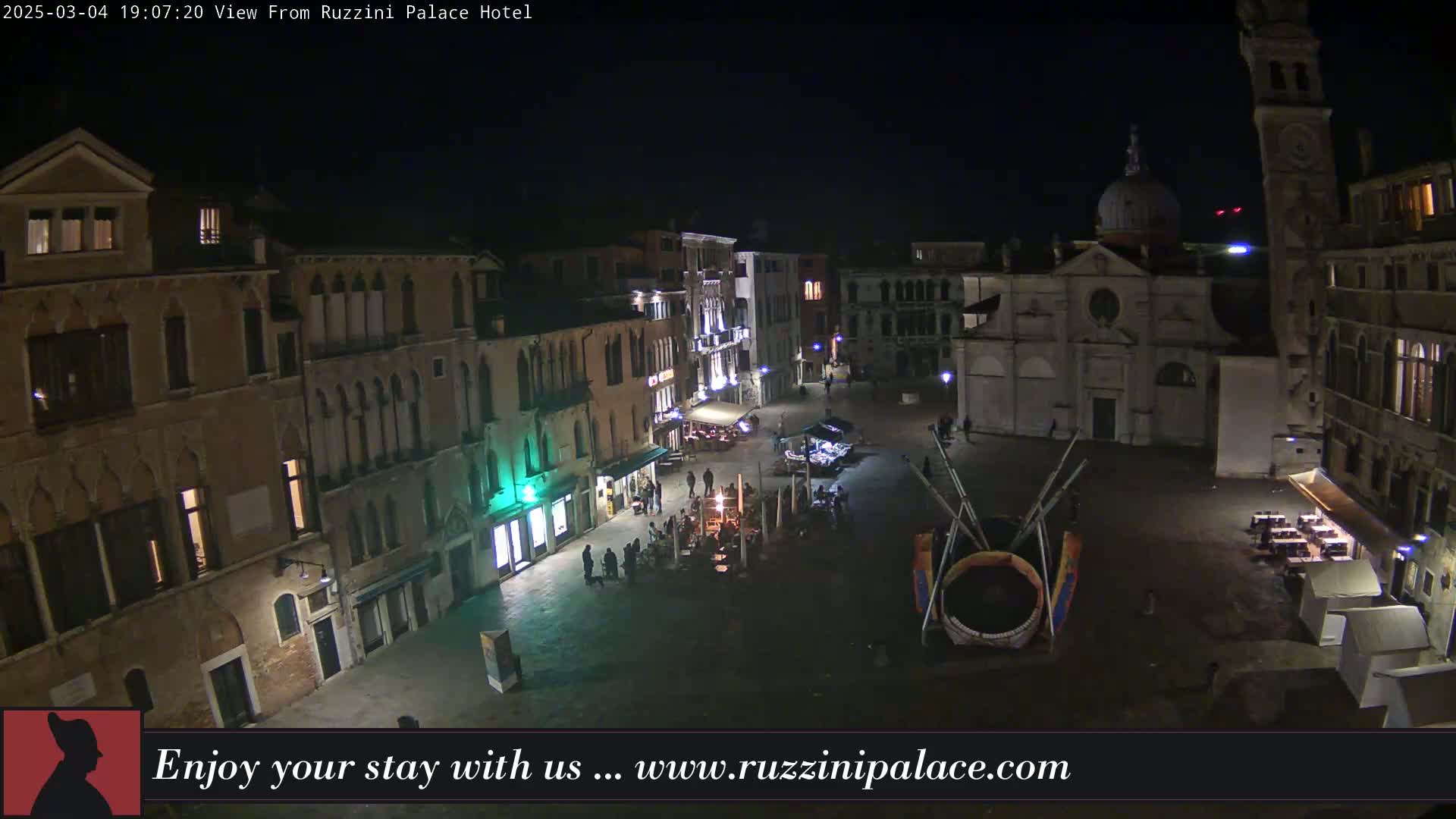 A nighttime aerial view of a dimly lit European plaza surrounded by old buildings, with a few people visible and some outdoor seating areas.