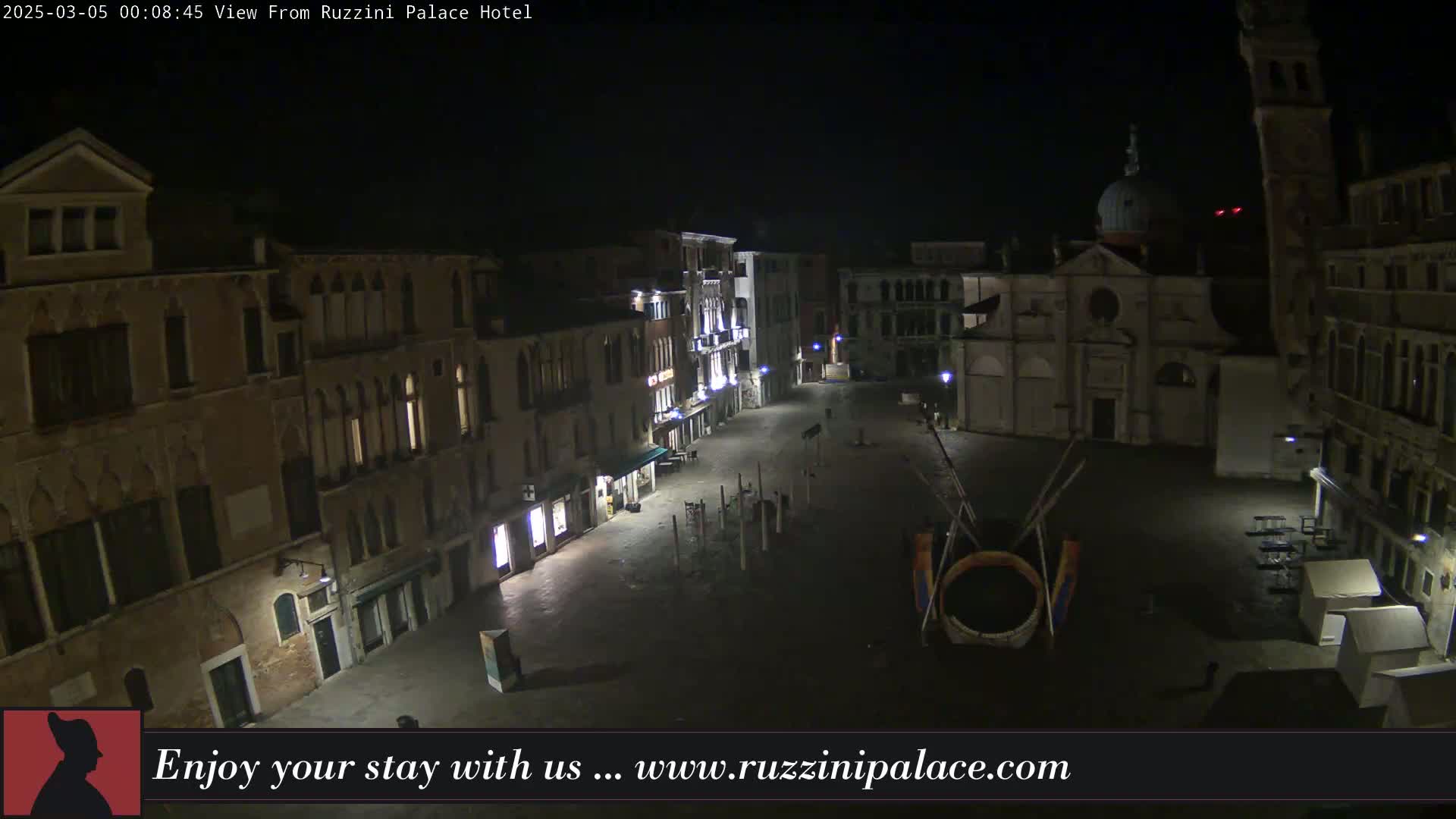 A nighttime, high-angle view of a mostly empty town square in Venice, Italy, surrounded by old buildings and a church.