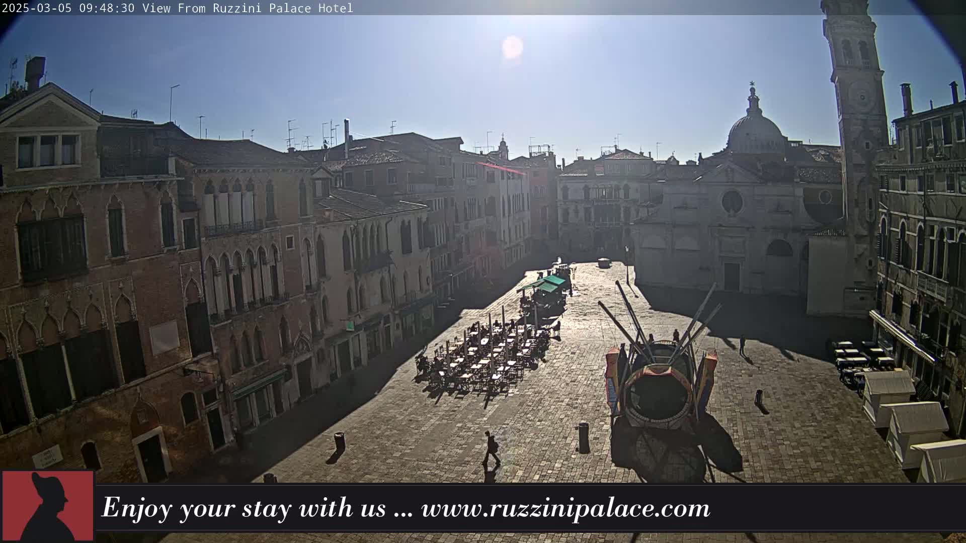A sunny day reveals a large, paved square in Venice, Italy, surrounded by historic buildings, with a few people and outdoor seating visible.
