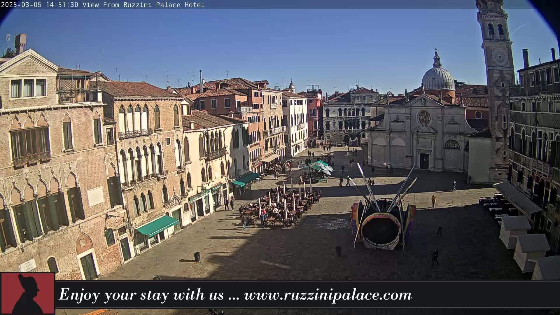 A sunny Venetian piazza with several multi-story buildings surrounding it, people walking and sitting at outdoor tables, and a large carnival-style ride in the center.