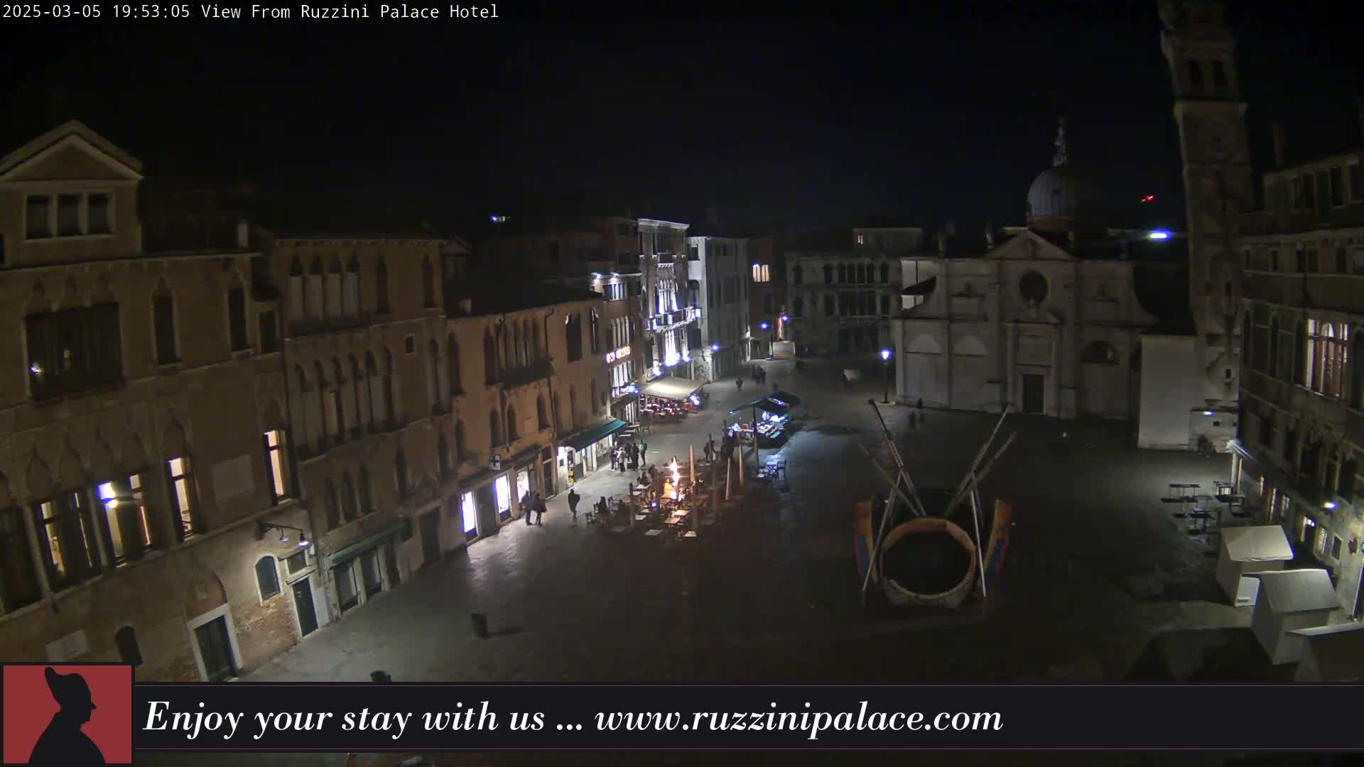 A nighttime view of a European town square, showing several buildings and a few people, under a dark sky.