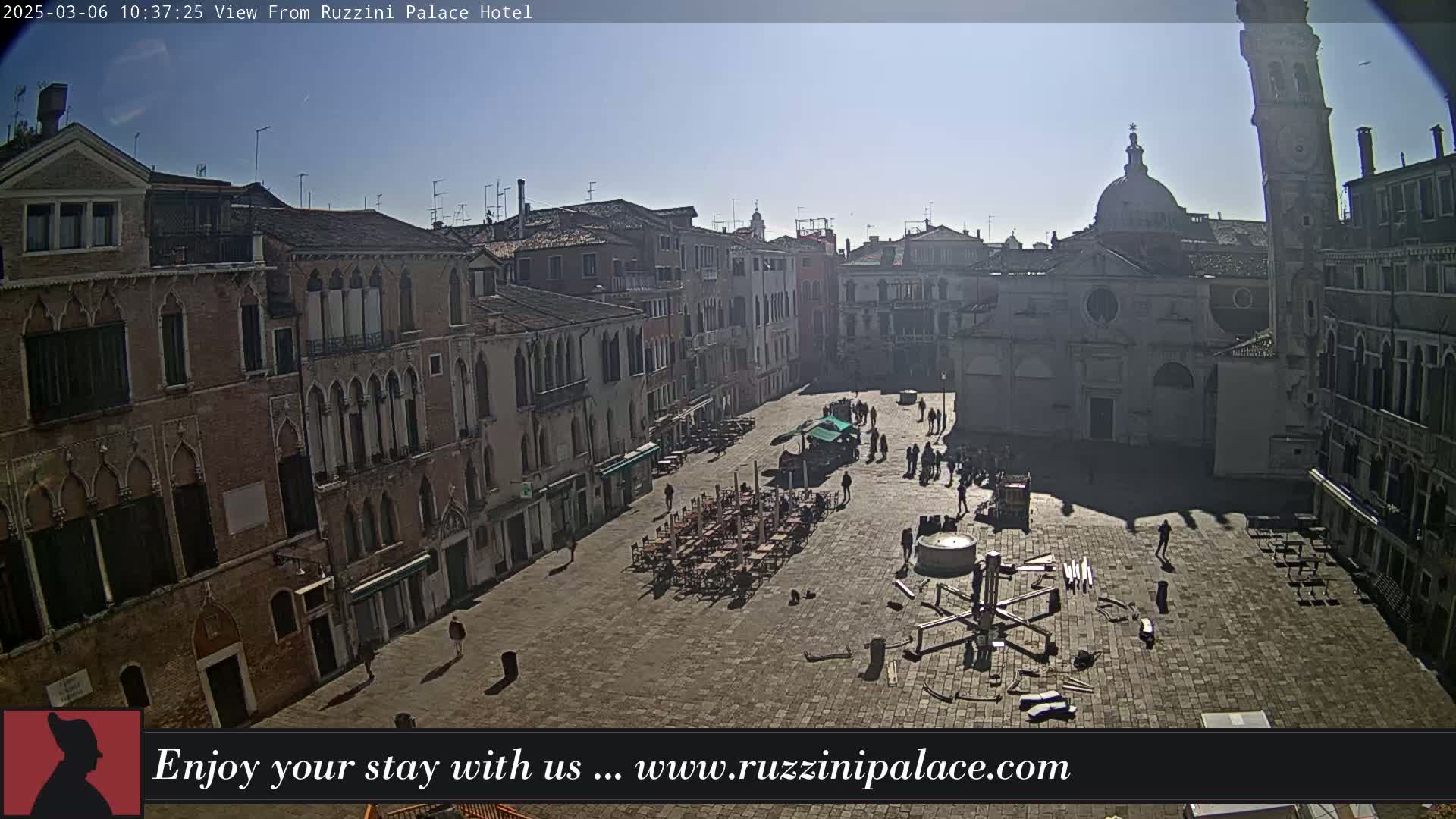 A sunny day reveals a large, paved square in Venice, surrounded by aged buildings, with a few people walking and some outdoor seating.