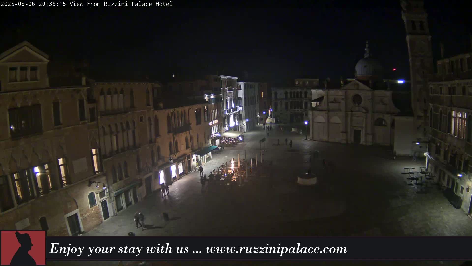 A nighttime, aerial view of a dimly lit European-style square, surrounded by aged buildings, with a few people walking around.
