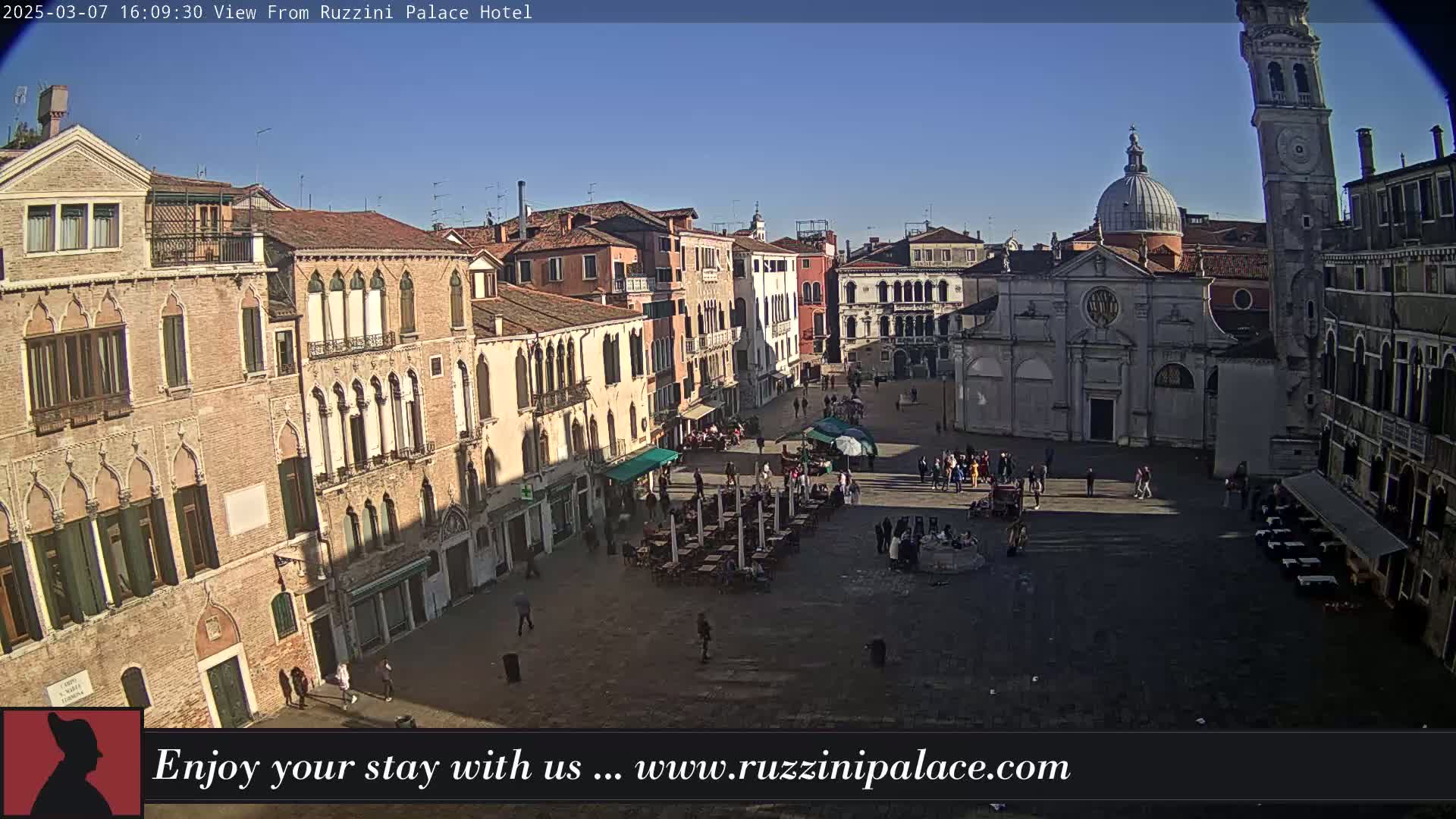 A sunny view of a large, paved square in Venice, Italy, surrounded by historic buildings, with people scattered throughout the square and outdoor seating at several cafes.