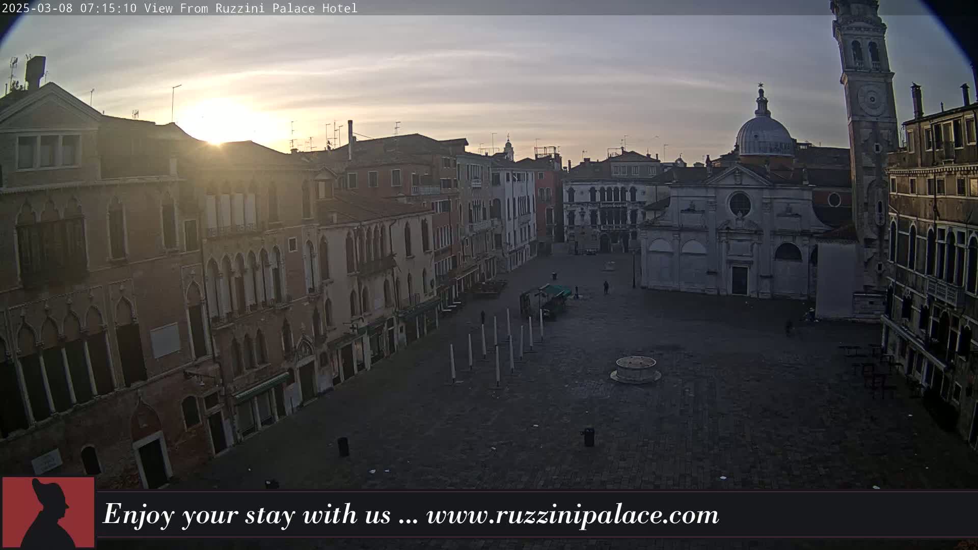 A sunrise illuminates a large, mostly empty square in Venice, surrounded by old buildings, including a church and a tall tower.