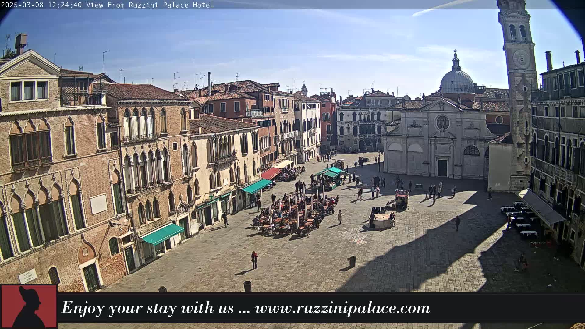 A sunny day reveals a large, paved square in Venice, Italy, surrounded by historic buildings, with several people and outdoor seating areas visible.